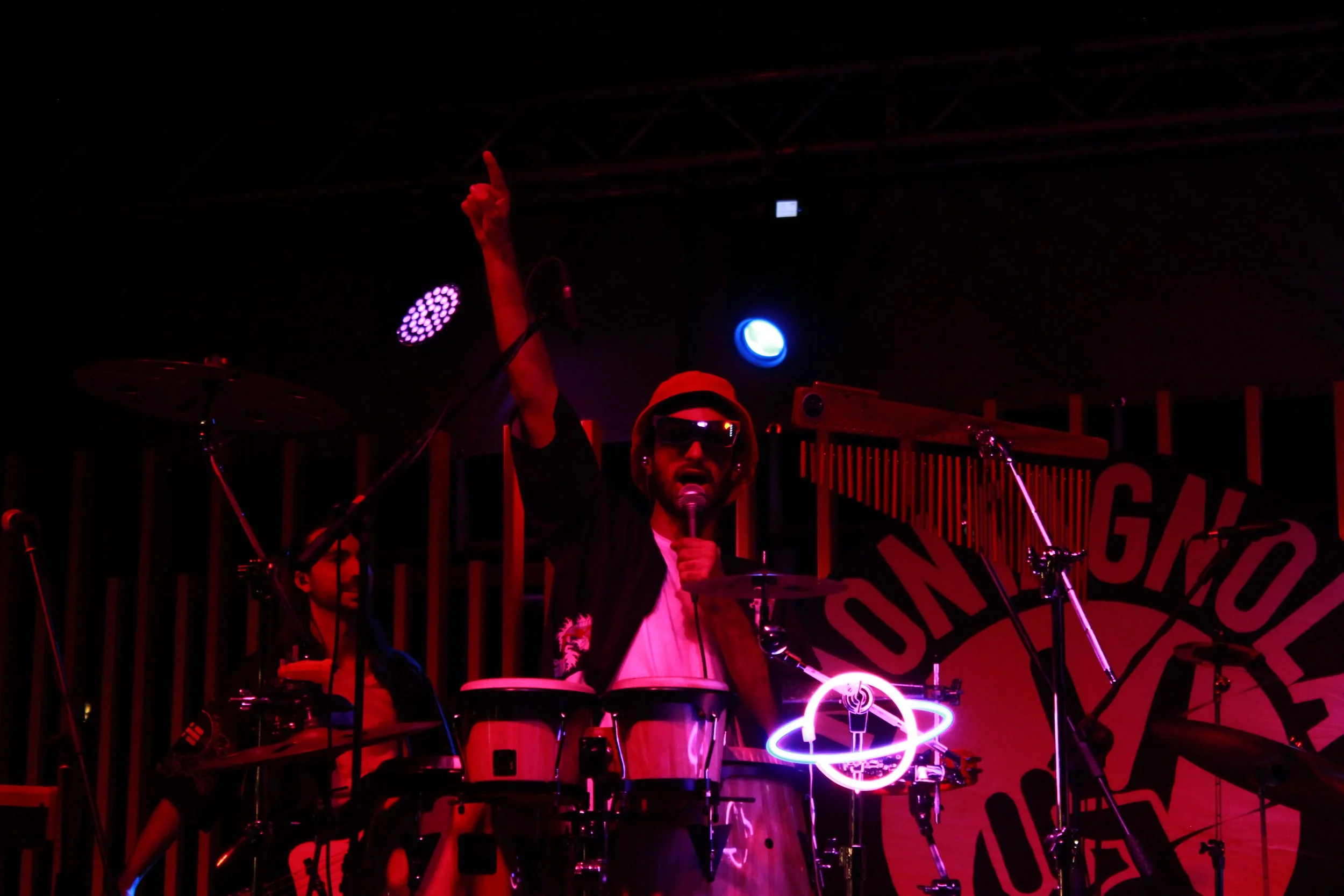 A person wearing sunglasses and a hat, singing into a microphone, playing percussion instruments, on a dimly lit stage with colorful stage lights and a neon light display of a planet with rings.