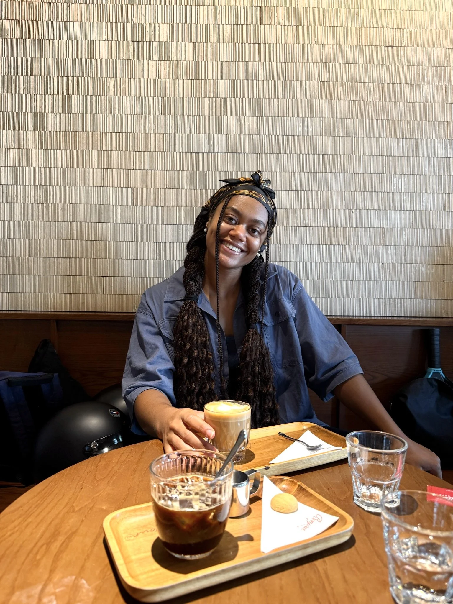 A smiling woman with braids, wearing a headband, sitting at a restaurant table, holding a coffee drink. The table has a tray with a coffee, a glass of water, and some small items.