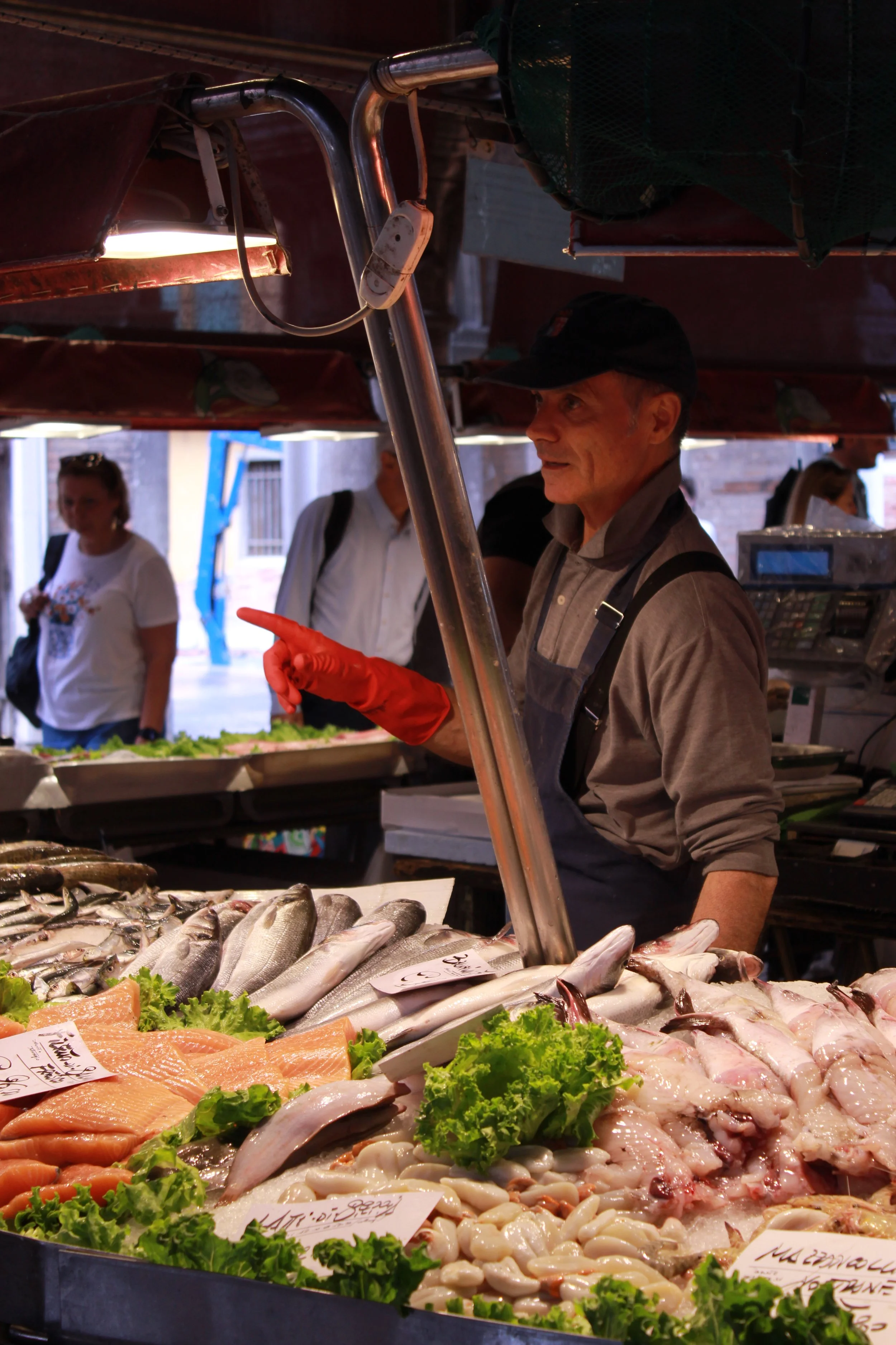 Market stall displaying fresh fish and seafood on ice, with a man wearing a black cap and gray shirt selecting seafood, surrounded by other shoppers.