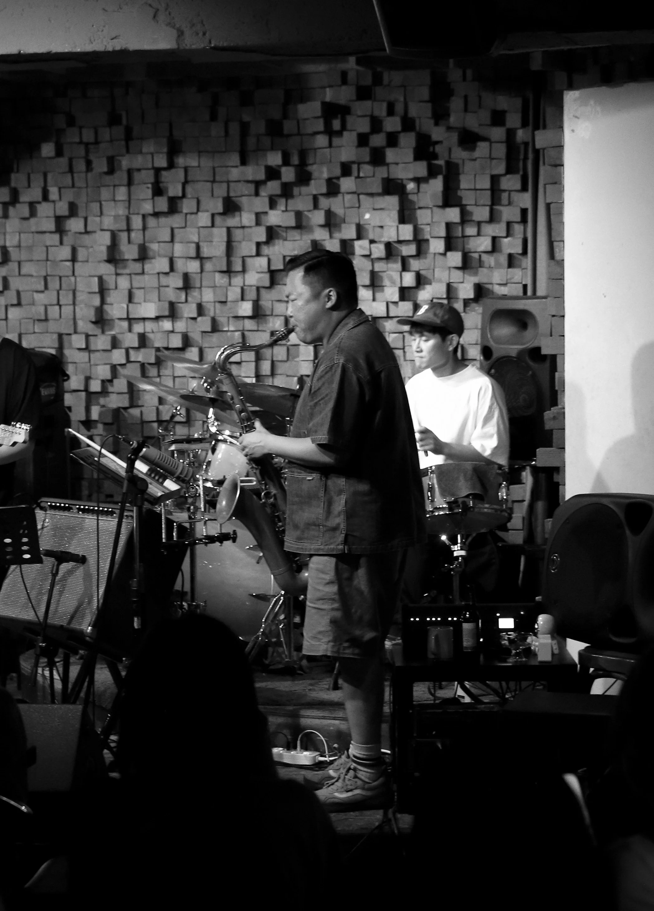 Black and white photo of musicians performing with electronic equipment, one playing a saxophone, in front of a textured brick wall.