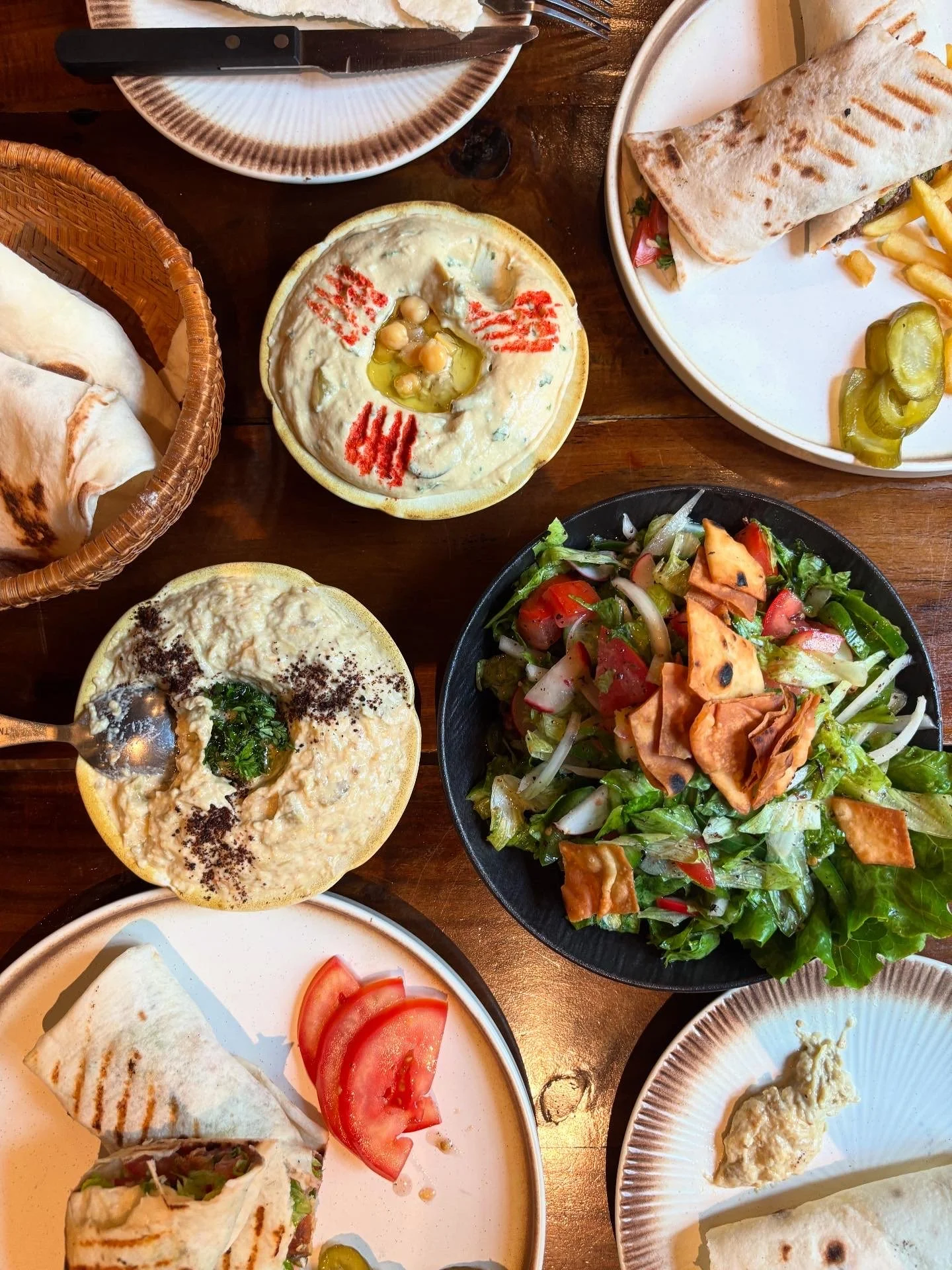 A variety of Lebanese dishes on plates and bowls, including hummus, a salad with lettuce and sliced vegetables, pita bread, and a wrap sandwich, on a wooden table.
