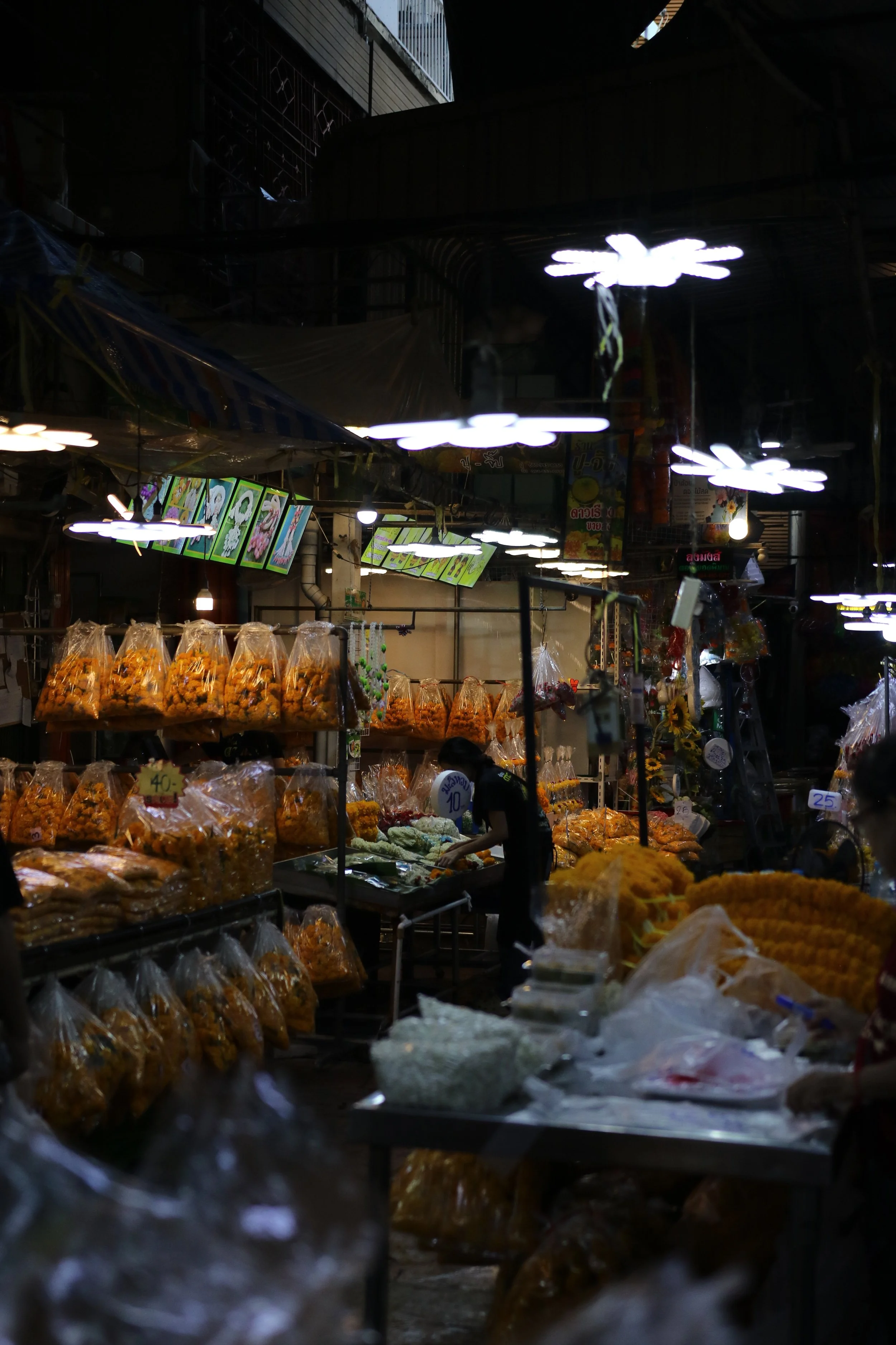 Indoor Thailand market stall with dried snacks and flowers, illuminated by hanging lights, with colorful menu boards overhead.