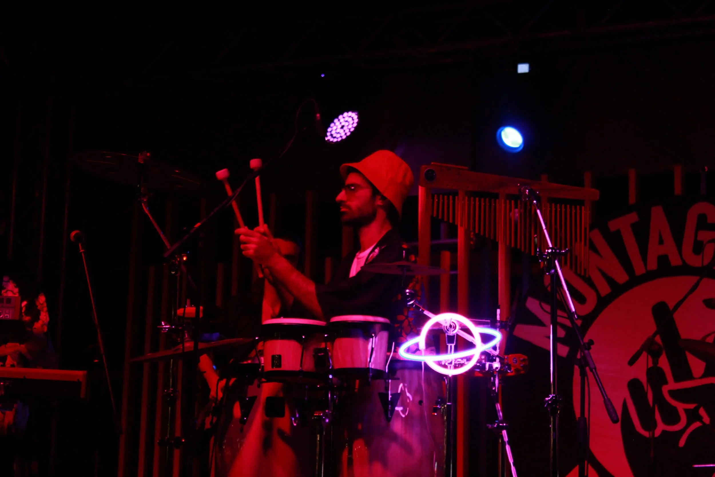A musician wearing a hat and glasses playing percussion instruments on stage, illuminated by colorful stage lights.