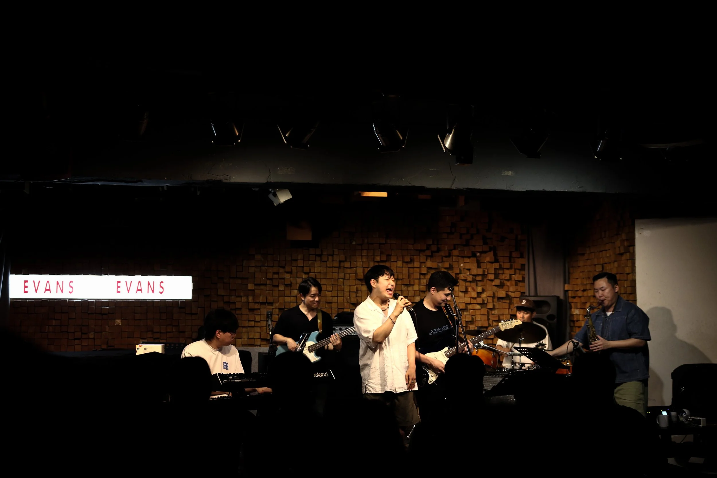 A band of musicians performing on stage with instruments including guitars, keyboard, drums, and saxophone. The background features a wooden wall with a sign that reads 'EVANS'. Several people are in the audience watching the performance.