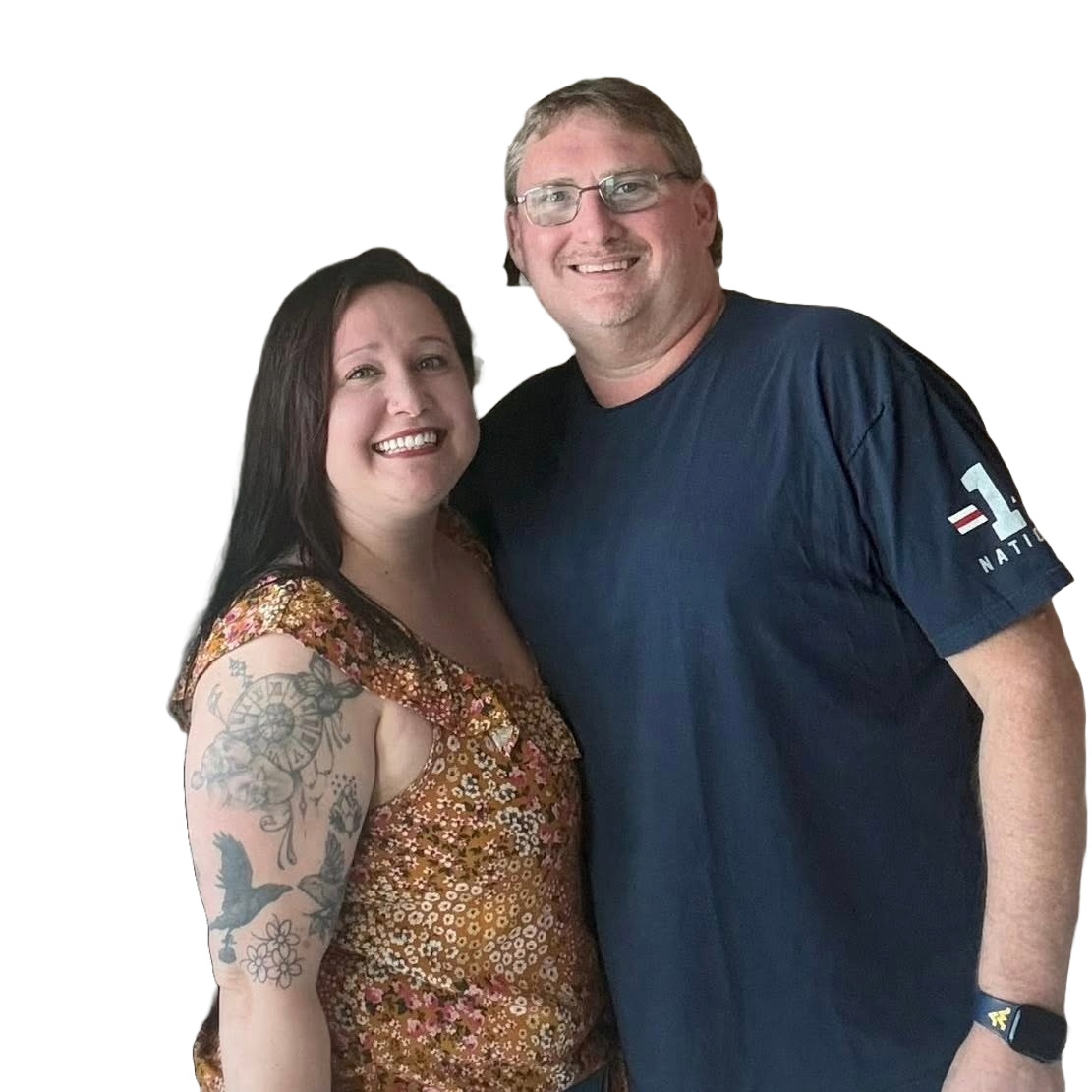 A smiling man and woman standing close together, the woman has dark hair and tattoos on her arm, and the man has glasses and light-colored hair, both wearing casual clothing.