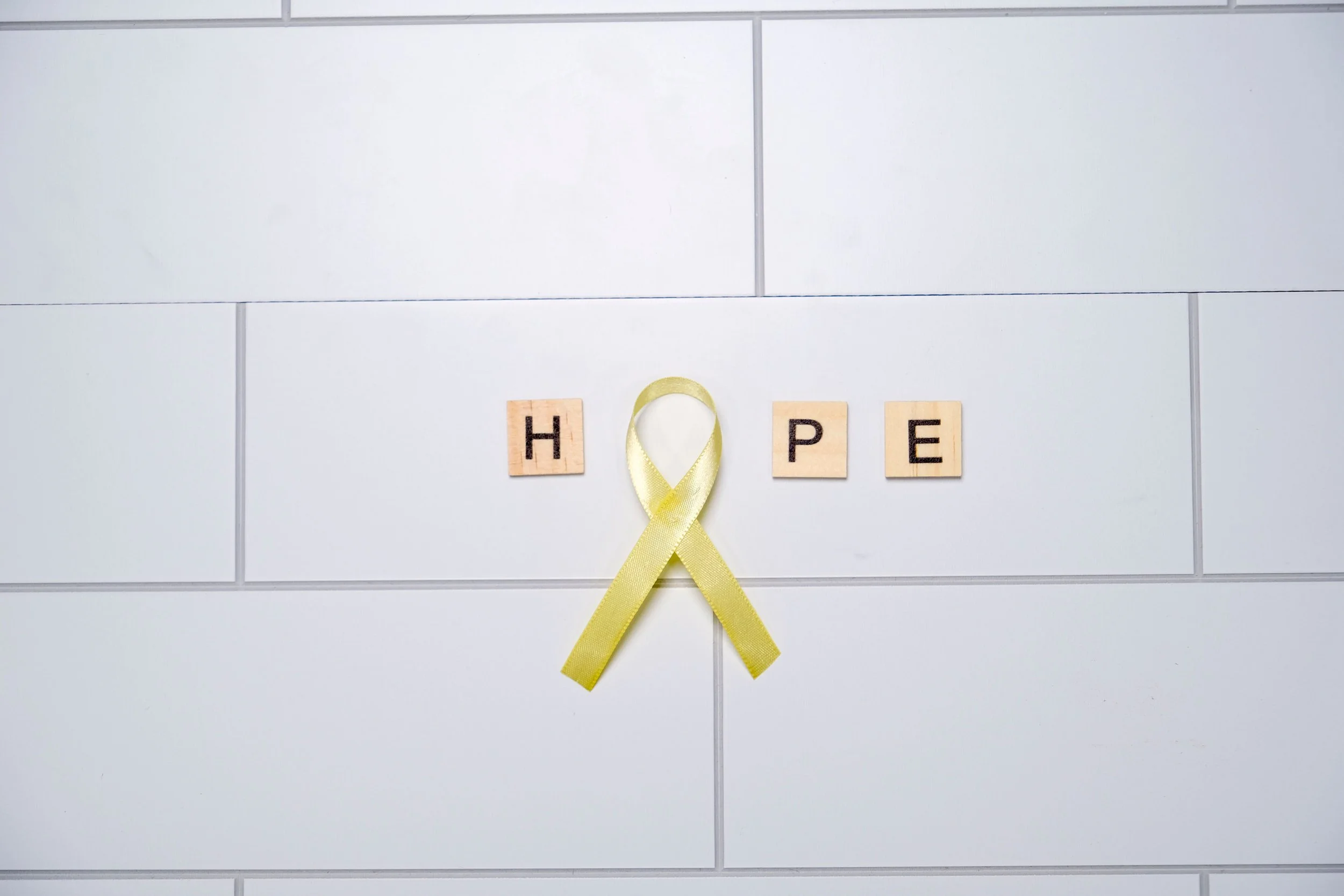 Scrabble tiles spelling 'HOPE' with a yellow awareness ribbon in front of a white tile wall.