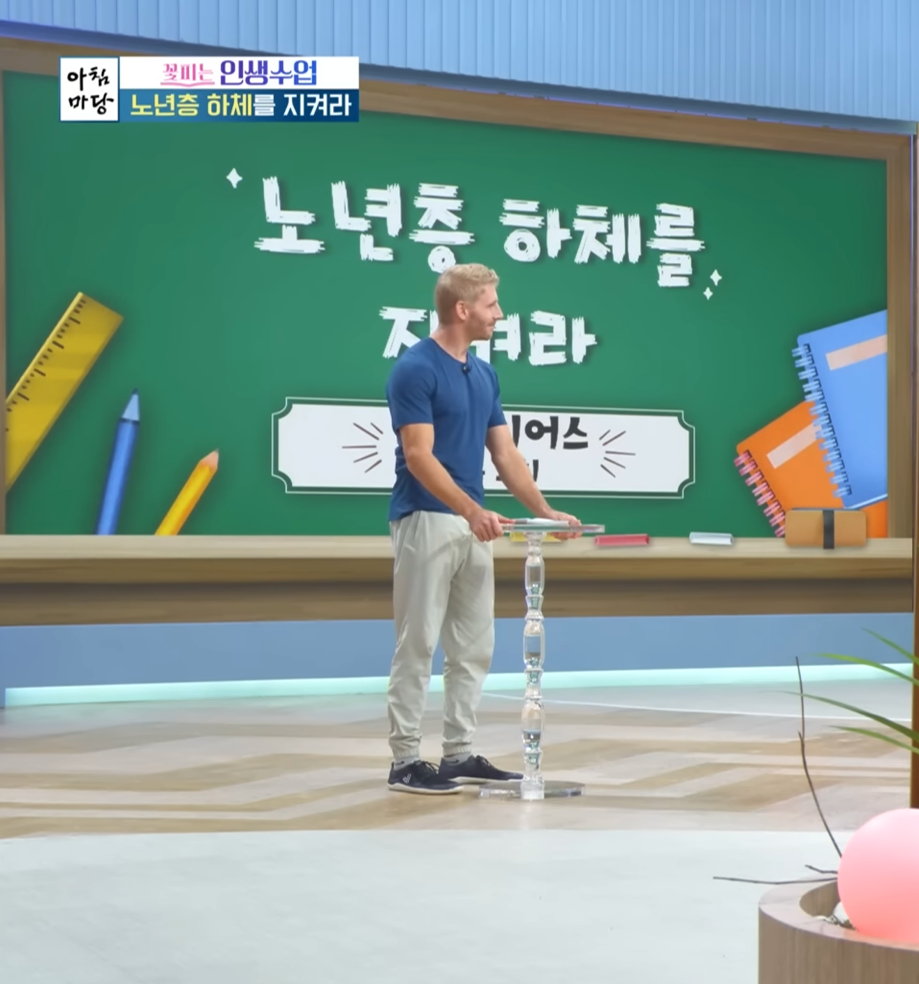 A man standing on a stage in front of a green chalkboard with Korean characters and school supplies illustrations, stacking clear glass cups.