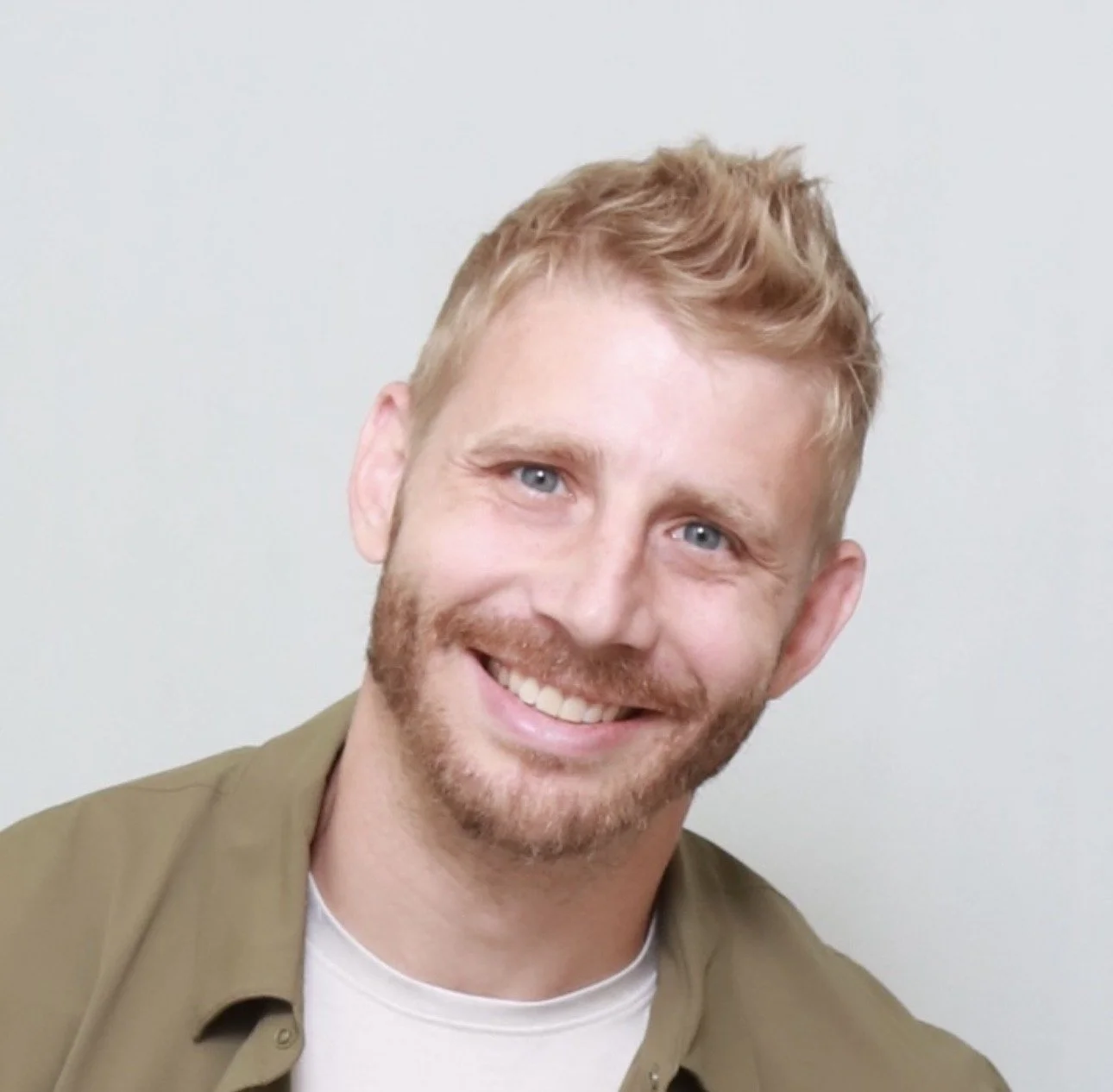 A smiling man with short, wavy blond hair, a beard, and blue eyes, wearing a beige shirt over a white t-shirt, against a plain white background.