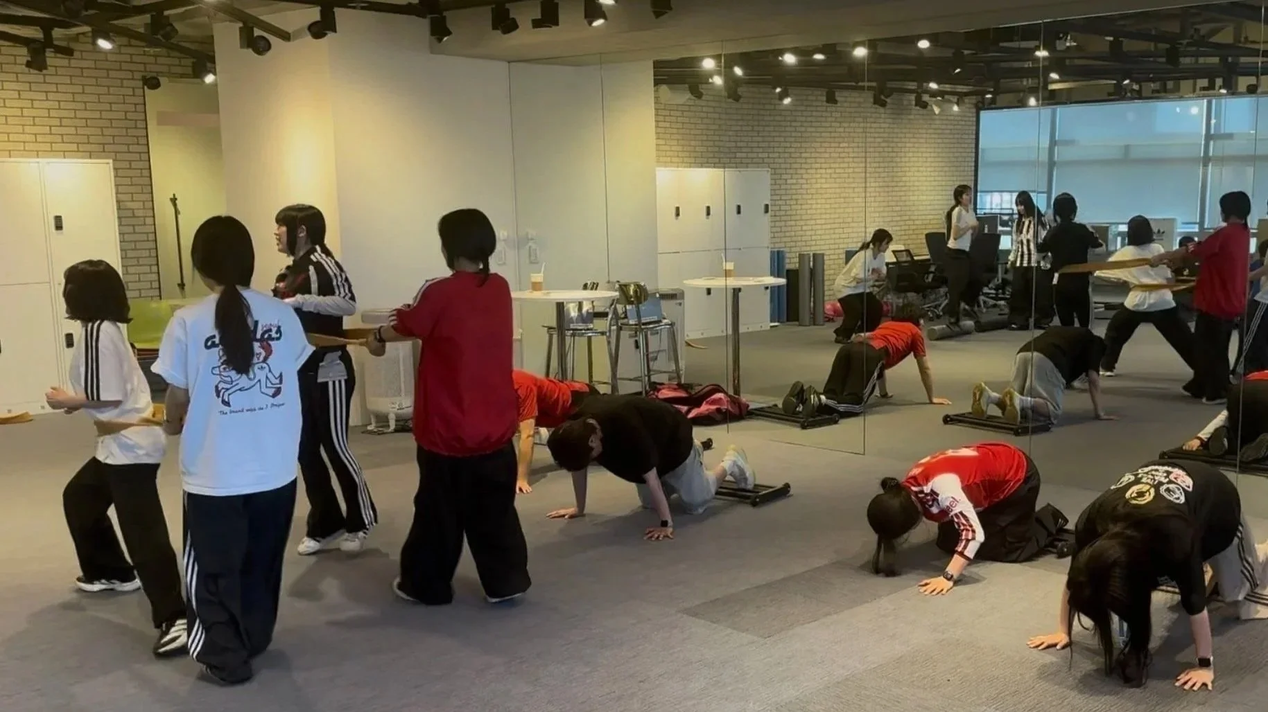 A group of people participating in a workout class in a corporate setting at Adidas headquarters in Seoul. The room has a mirror wall, lockers, and gym equipment.