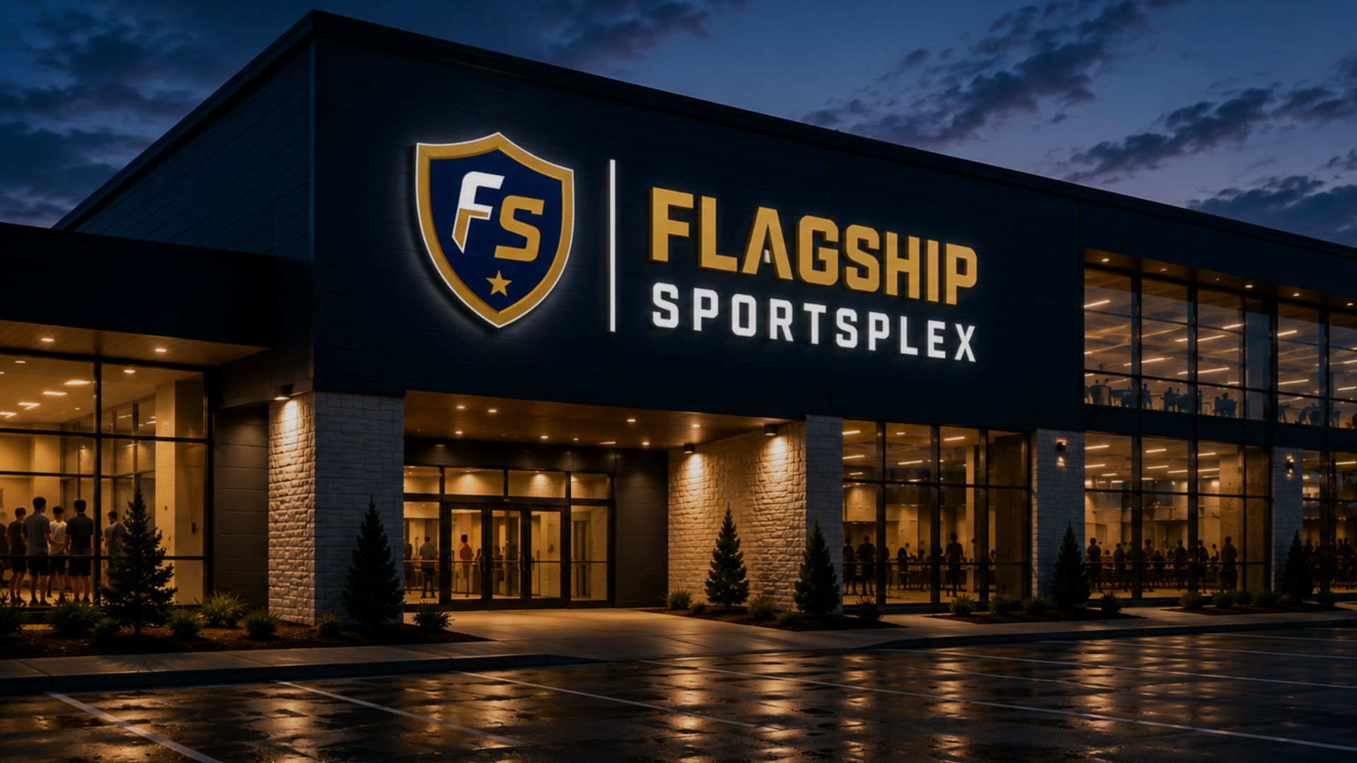 Night view of the Flagship Sportsplex building with illuminated sign, large glass windows revealing people inside, and a parking lot with wet pavement reflecting lights.