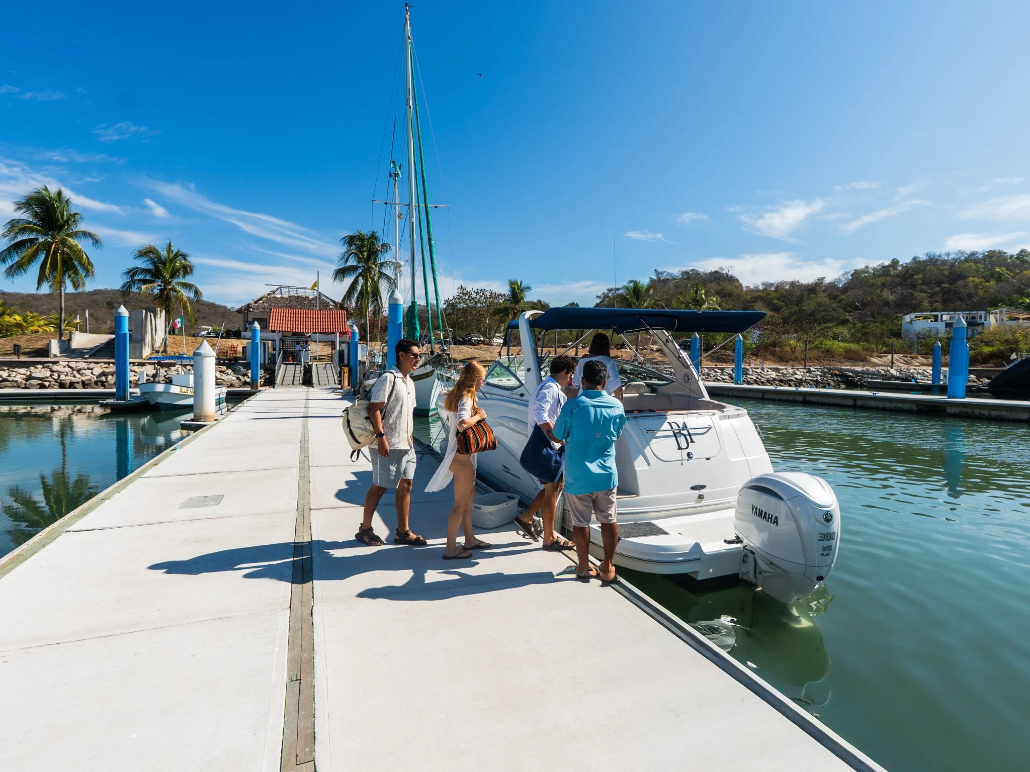 Personas abordando un yate en un muelle en una zona costera con palmeras y vegetación, cielo despejado y en un día soleado.