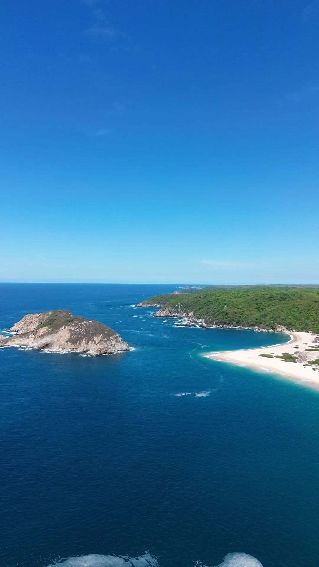 Vista aérea de una playa con arena blanca, aguas azules y colinas verdes en el fondo.