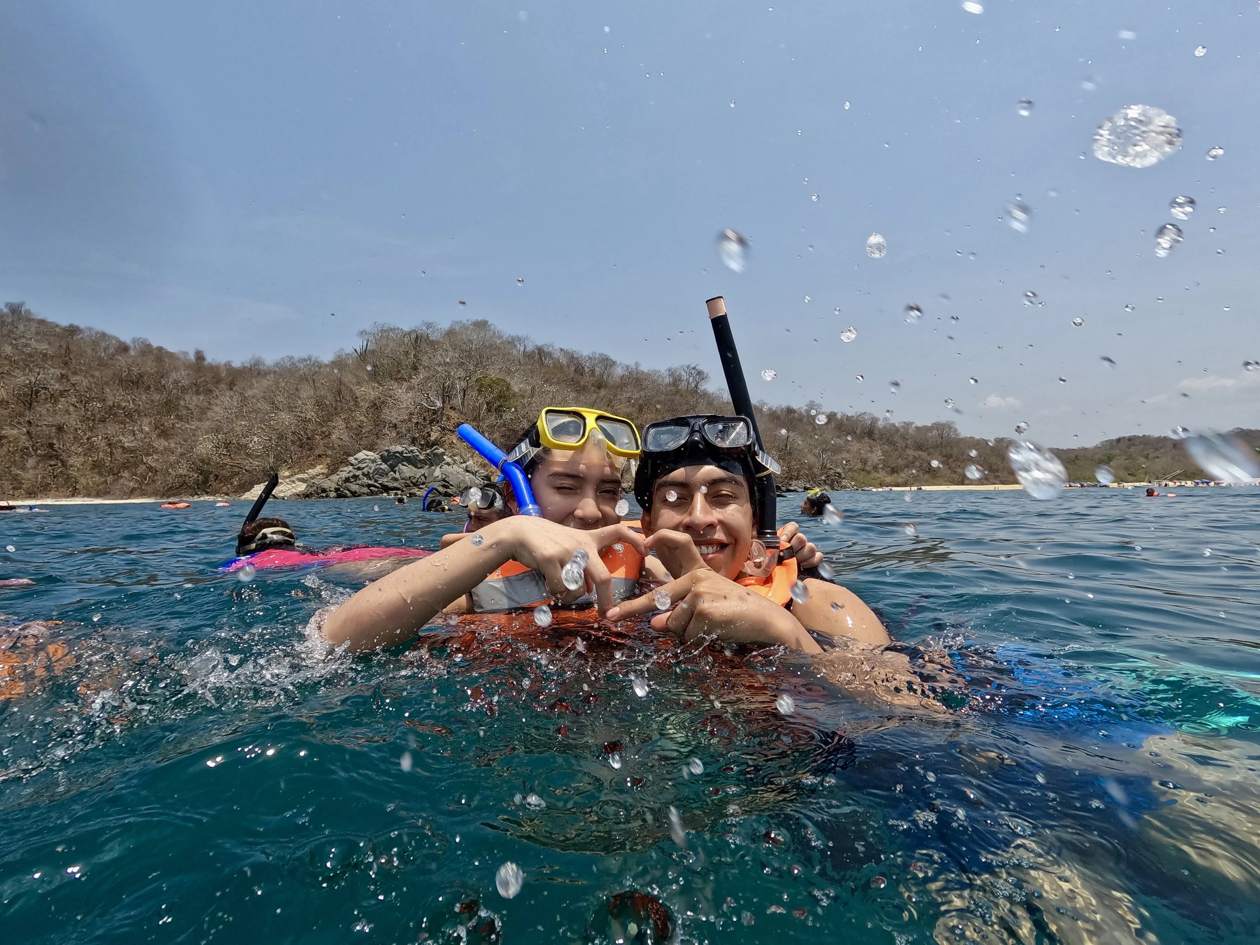 Dos personas con gafas de snorkeling y trajes de baño en el agua, haciendo un corazón con las manos mientras nadan en un río o mar, con fondo de colinas y árboles secos.