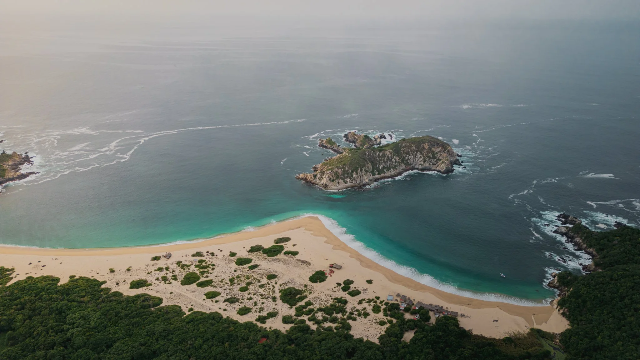 Una playa arenosa con aguas turquesa, rodeada de vegetación verde y formaciones rocosas en el océano