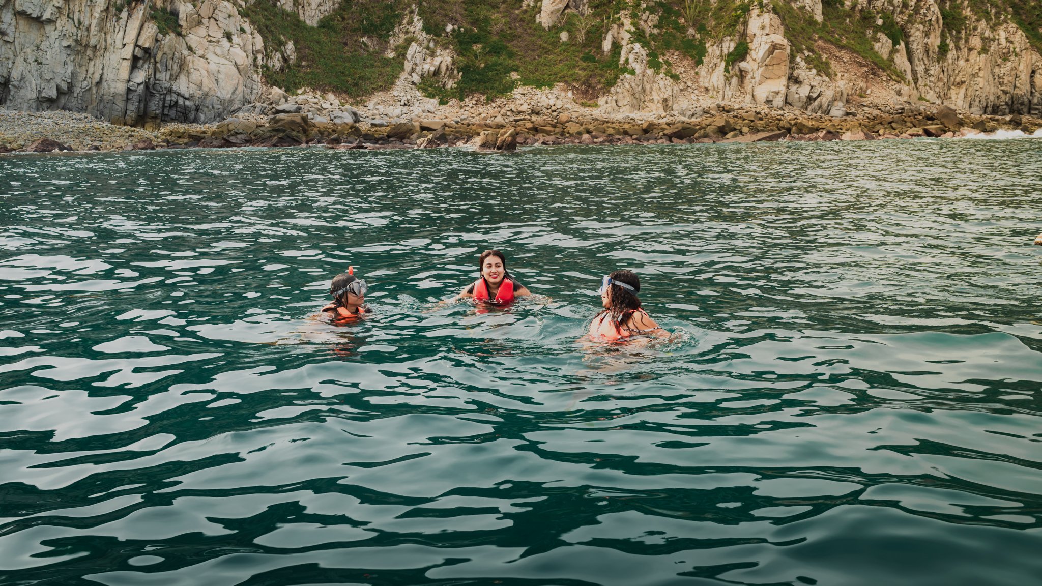 Tres personas nadando en el mar cerca de una costa con acantilados y rocas de fondo.