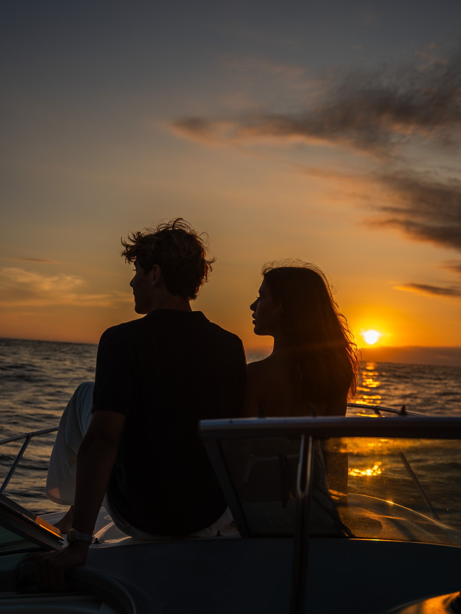 Dos personas, un hombre y una mujer, sentadas en un barco durante un atardecer en el mar, con el sol cercano al horizonte y el cielo con nubes.