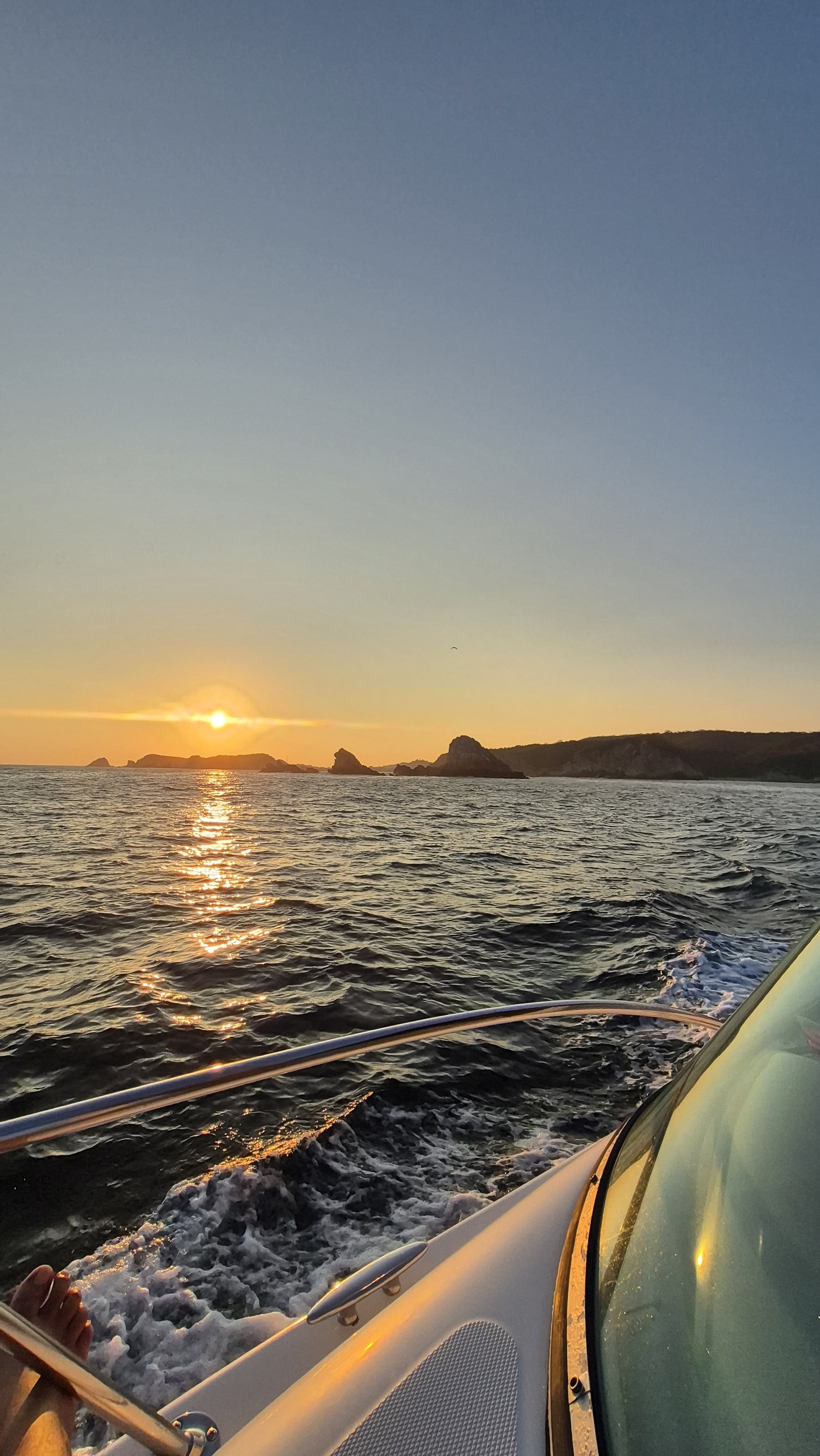 Atardecer en el mar visto desde una embarcación, con rocas en la línea del horizonte y olas en el agua.
