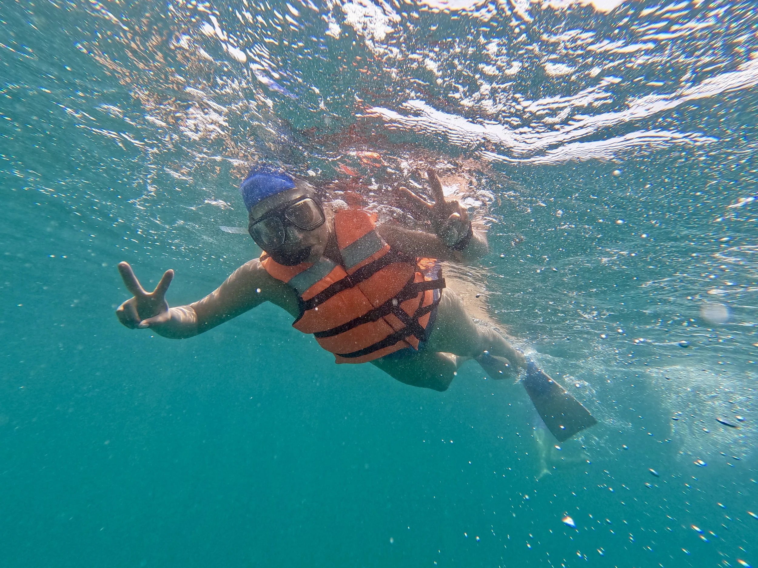 Persona haciendo snorkel en el mar, usando gafas y chaleco salvavidas, señalando con la mano y haciendo señal de paz.