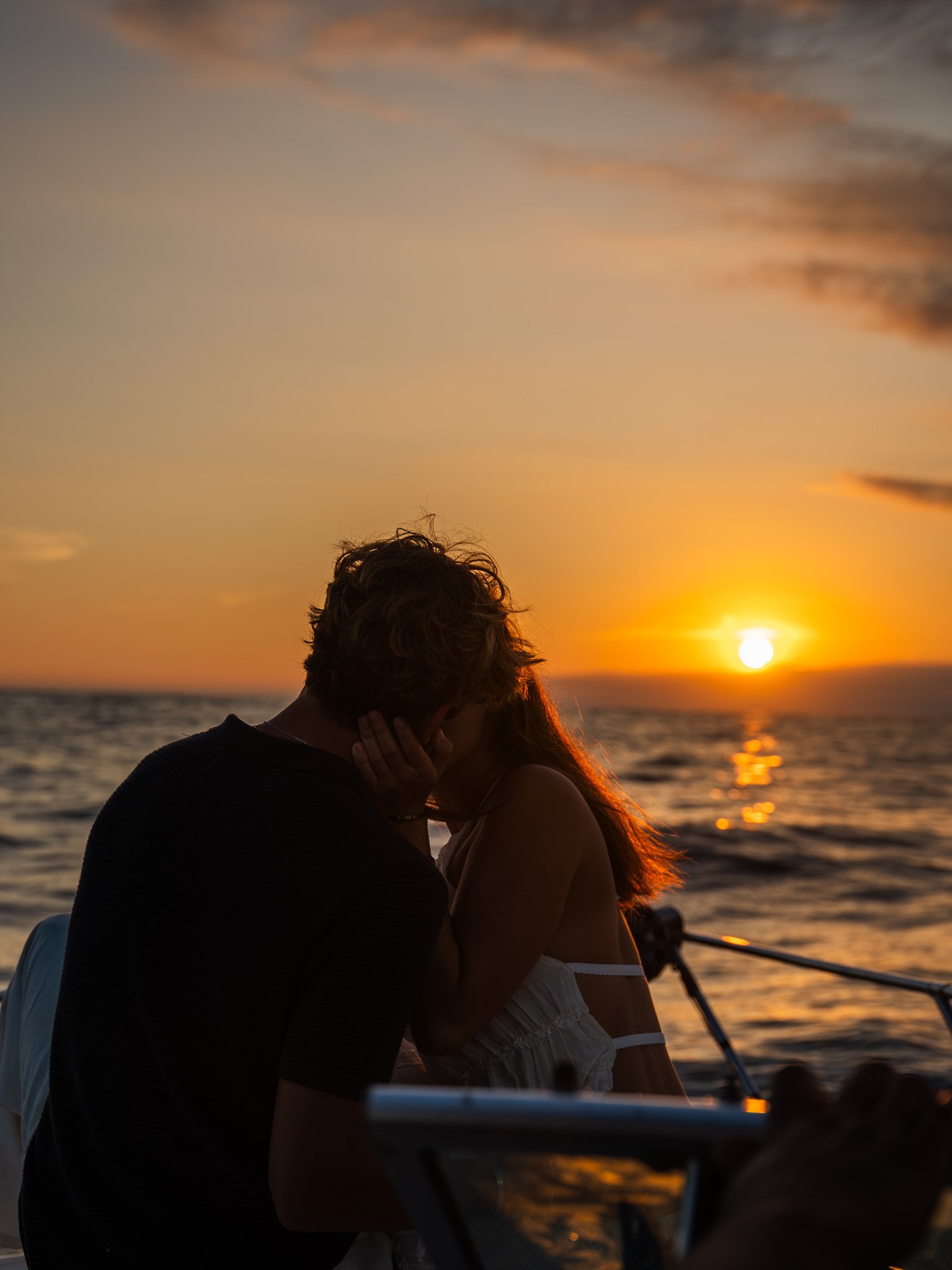 Una pareja se abraza y se besa en la orilla del mar durante una puesta de sol, con el cielo teñido de tonos anaranjados y el sol reflejado en el agua.