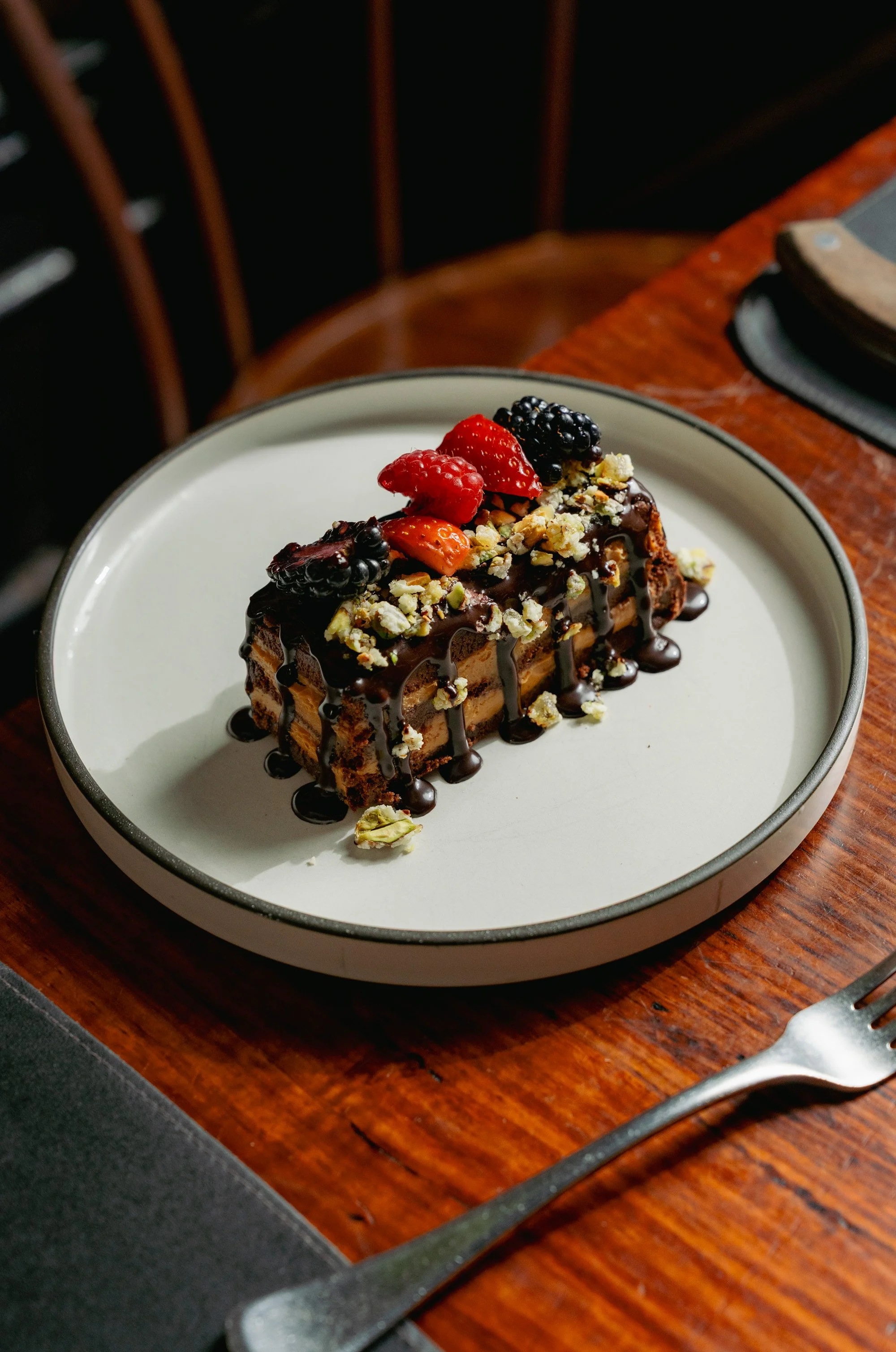 A slice of chocolate cake topped with mixed berries, chopped nuts, and drizzled with chocolate sauce on a white plate.