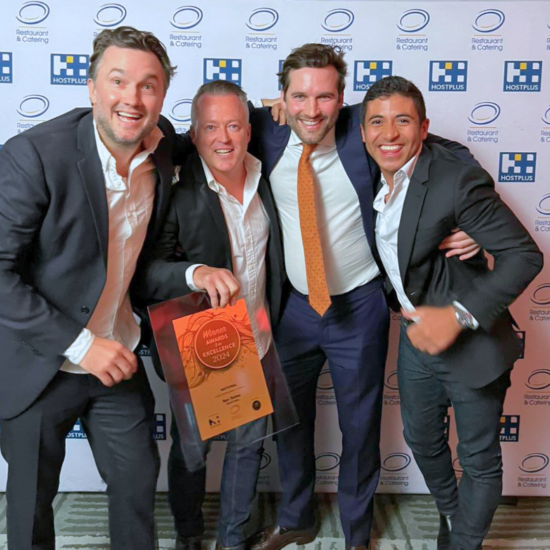 Four men in suits smiling and posing together at an award ceremony, one holding a plaque that reads "Winner Awards Excellence 2024". They stand in front of a backdrop with logos for "HostPlus" and "Restaurant & Catering."