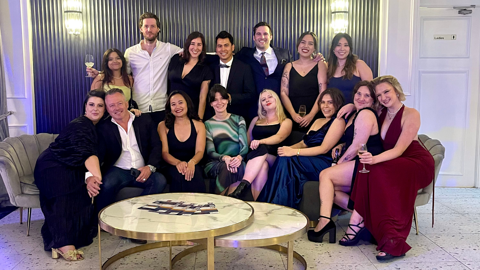 Group of people dressed in formal attire at a party, smiling and holding drinks, posing for a photo in a stylish lounge with a dark striped wall and a door labeled 'Ladies'
