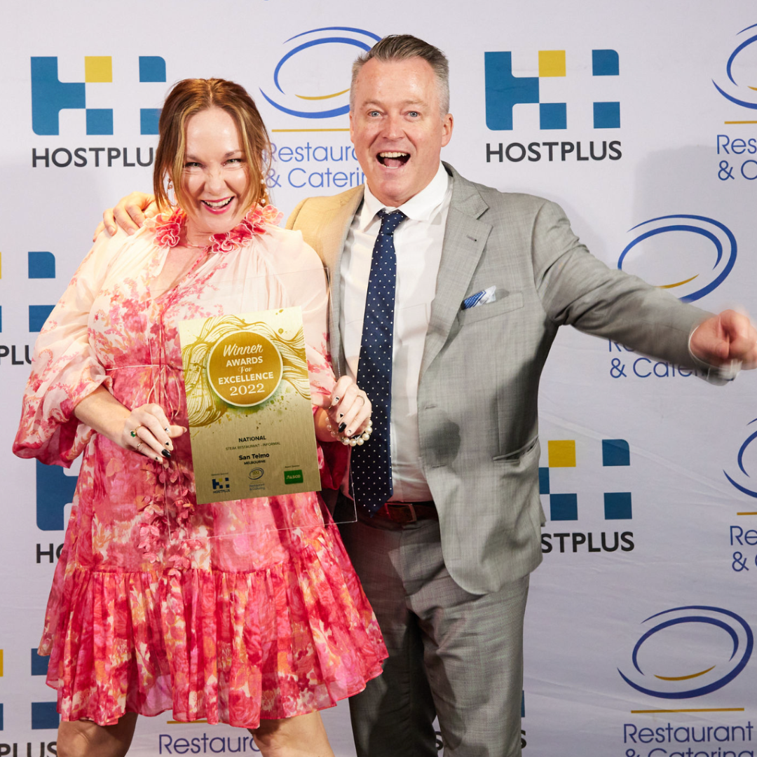 A man and woman in formal attire celebrating, holding an award certificate that reads 'Winner Awards for Excellence 2022,' at a promotional event for Hostplus, Restaurant & Catering.