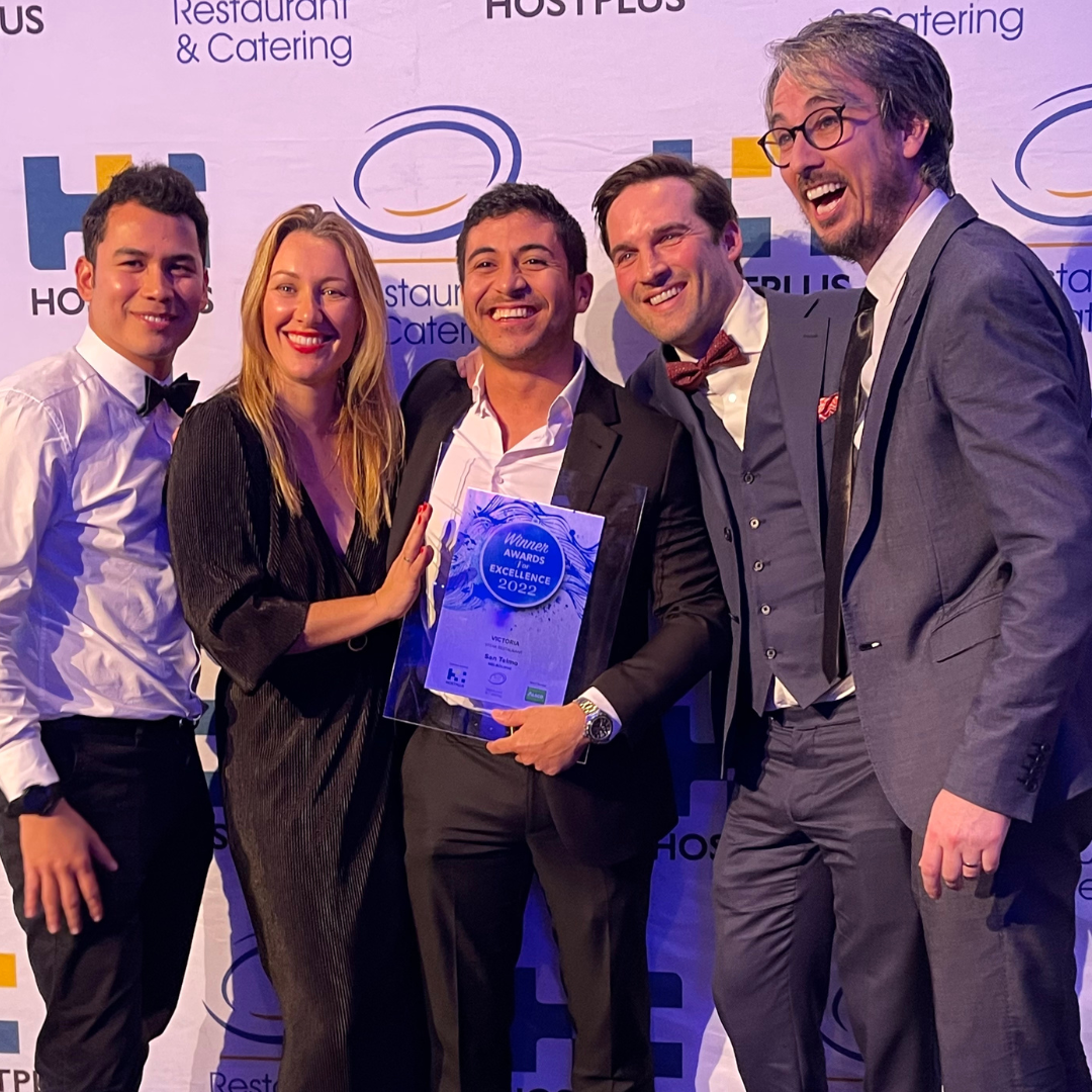 Group of five people in formal attire at an awards ceremony, holding a trophy, with a backdrop featuring logos and the words "Restaurant & Catering" and "Hostplus".