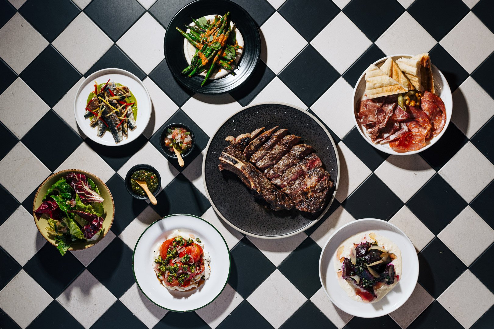 A variety of dishes including grilled steak, salads, grilled vegetables, cured meats, pita bread, and dips, arranged on a black and white checkered tiled surface.