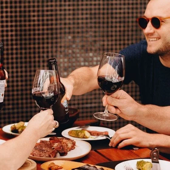 Two people clinking glasses of red wine at a dinner table with plates of food.
