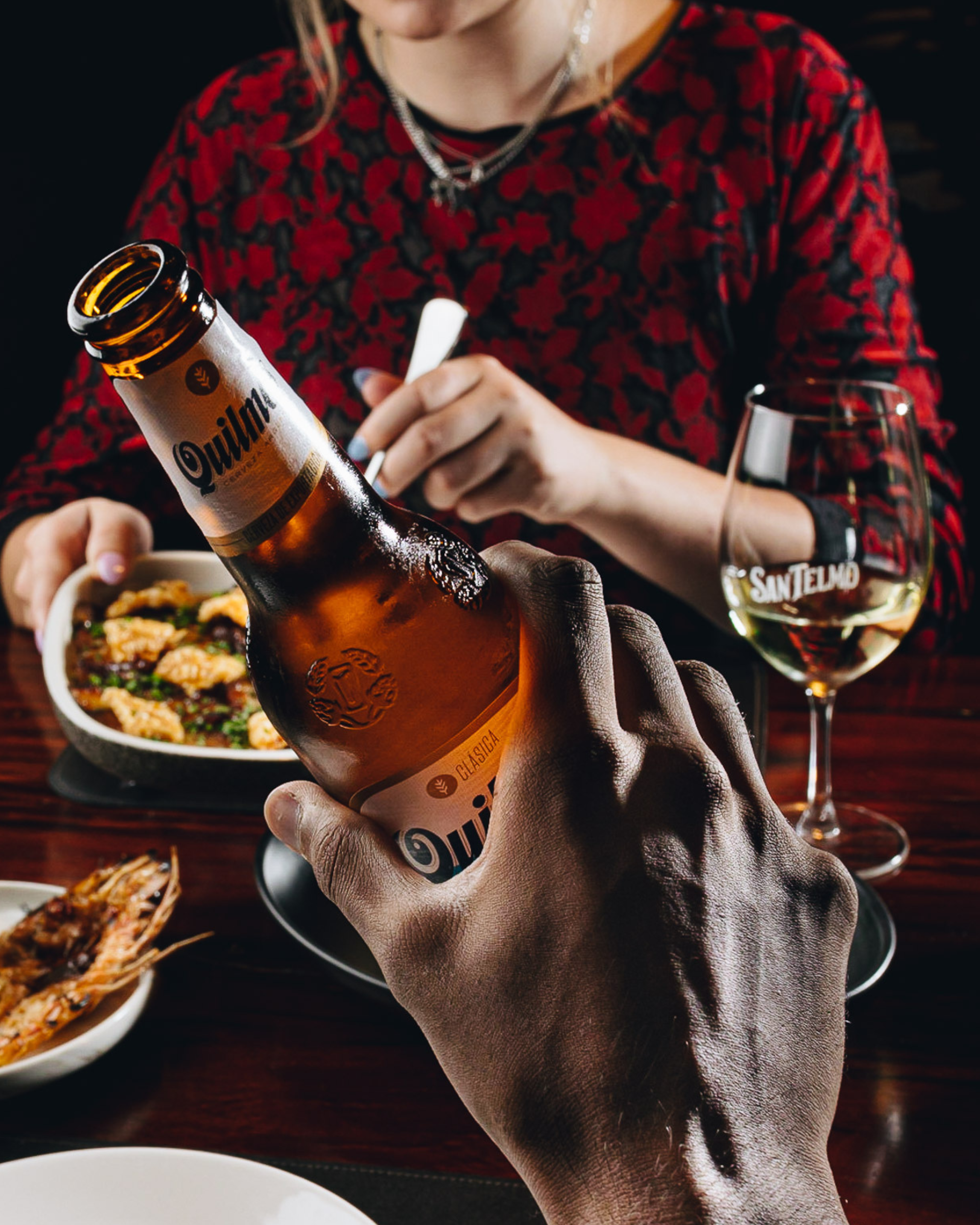 Close-up of a person holding a bottle of Quilmes beer with their hand, a glass of Sam Telmo beer, and a plate of food on a dark wooden table in the background. The person is wearing a red and black floral-patterned shirt and silver jewelry.