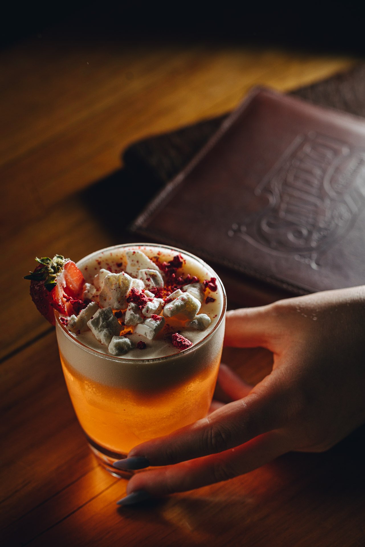 A hand holding a glass of beer topped with whipped cream, crushed red pepper, and a strawberry, with a chocolate bar in the background on a wooden surface.