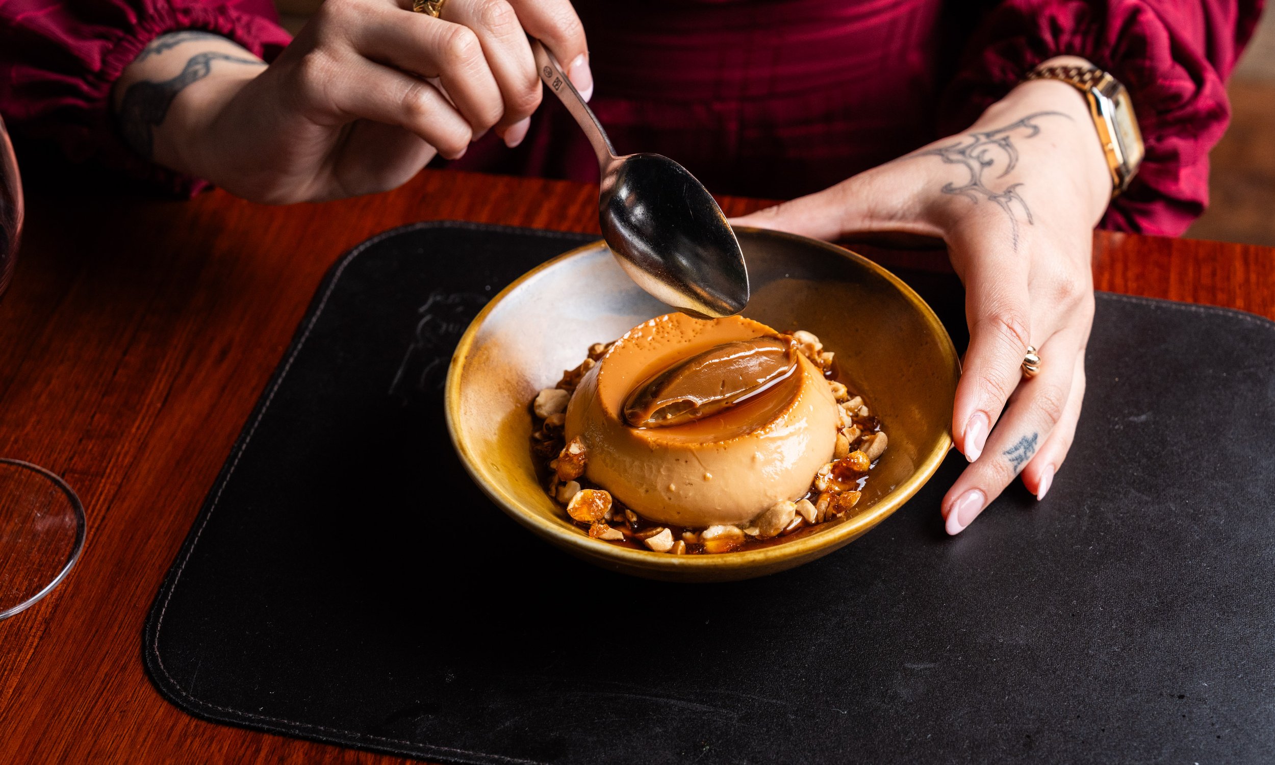 Person with tattoos on their hands and arms holding a spoon over a caramel flan dessert topped with caramel sauce and surrounded by chopped nuts on a yellow plate.