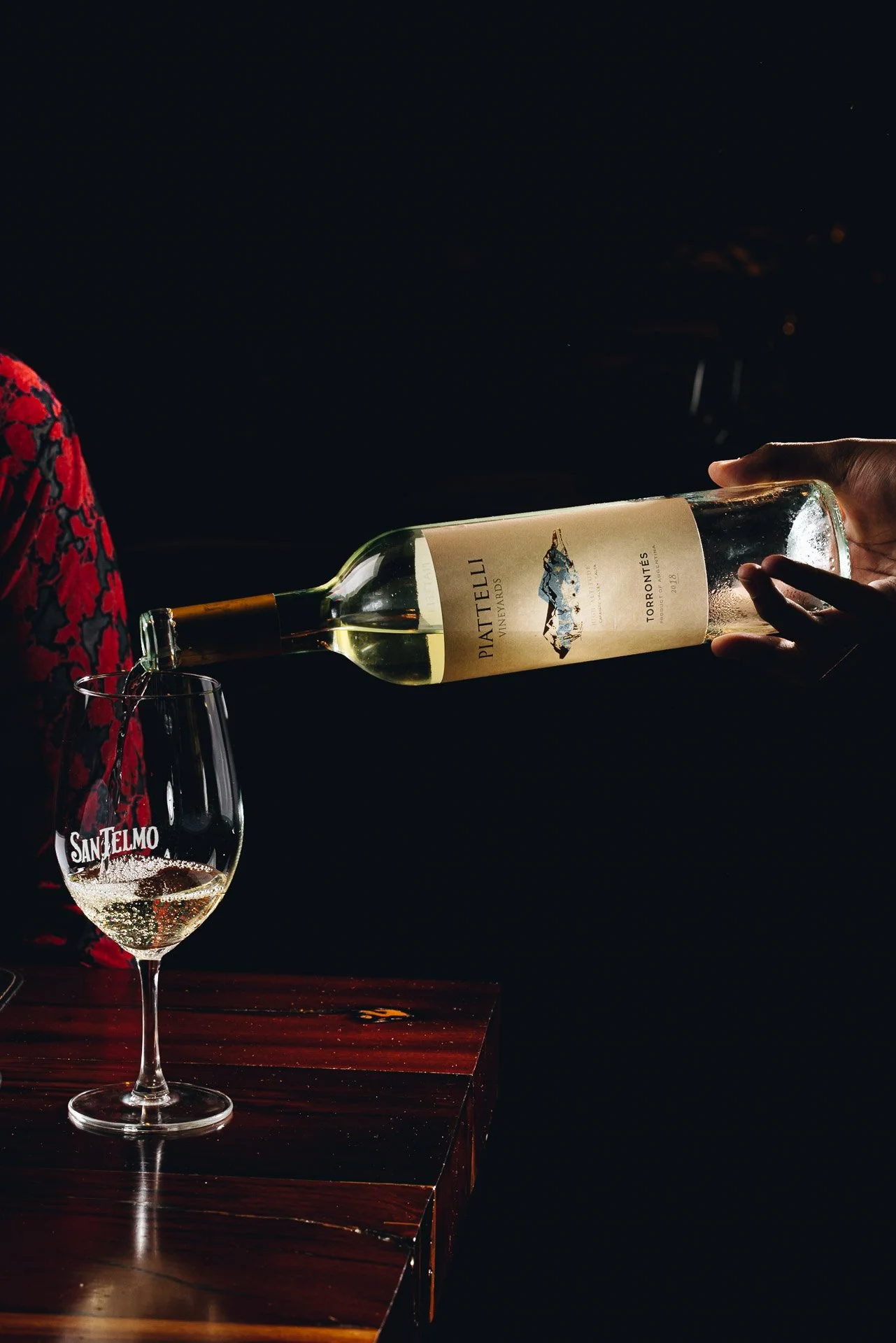 Person pouring white wine from a bottle into a wine glass set on a polished wooden surface against a dark background.