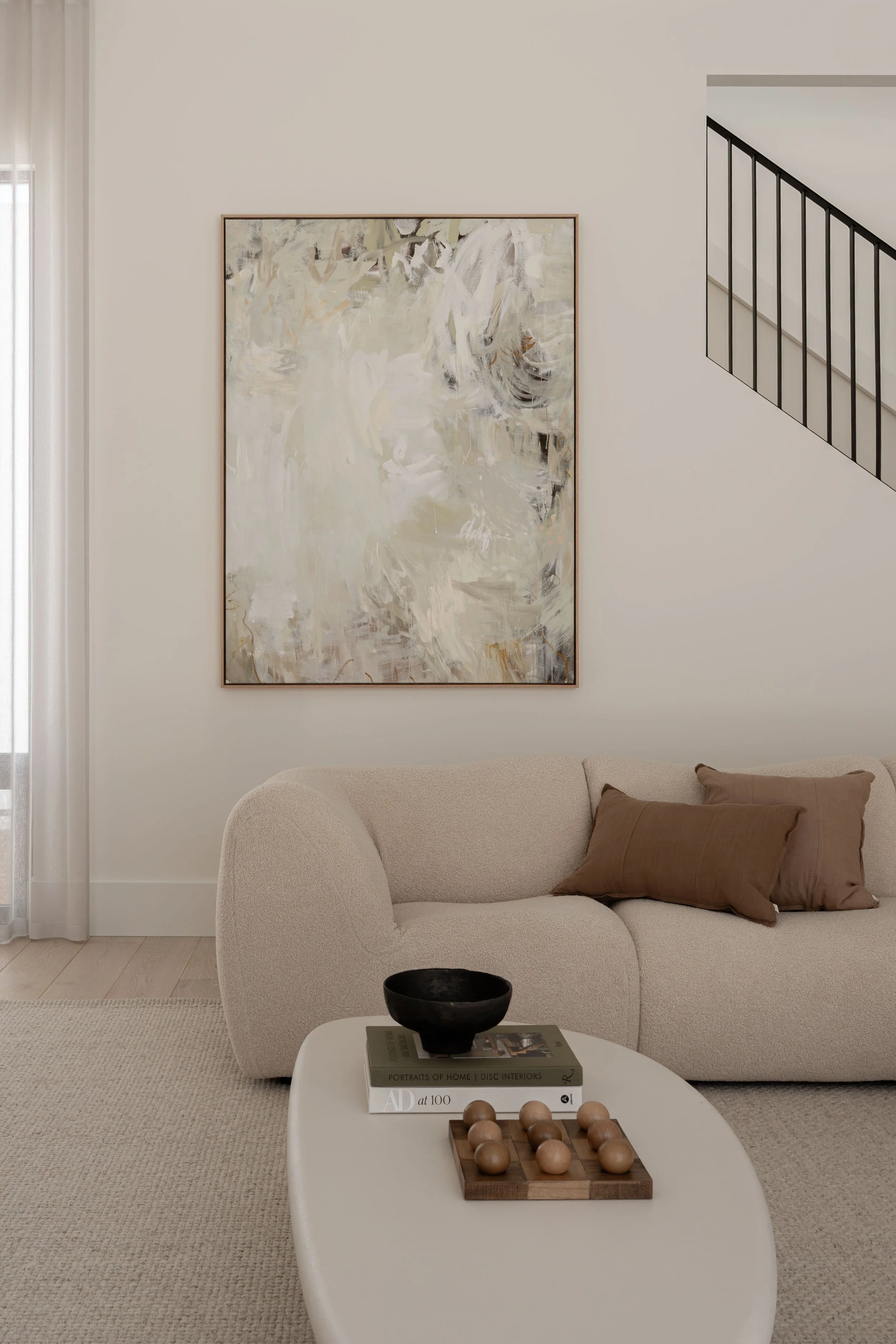 Living room with a beige sofa, brown pillow, abstract painting on the light-colored wall, black railing on the staircase, and a white coffee table with books, a black bowl, and wooden balls.