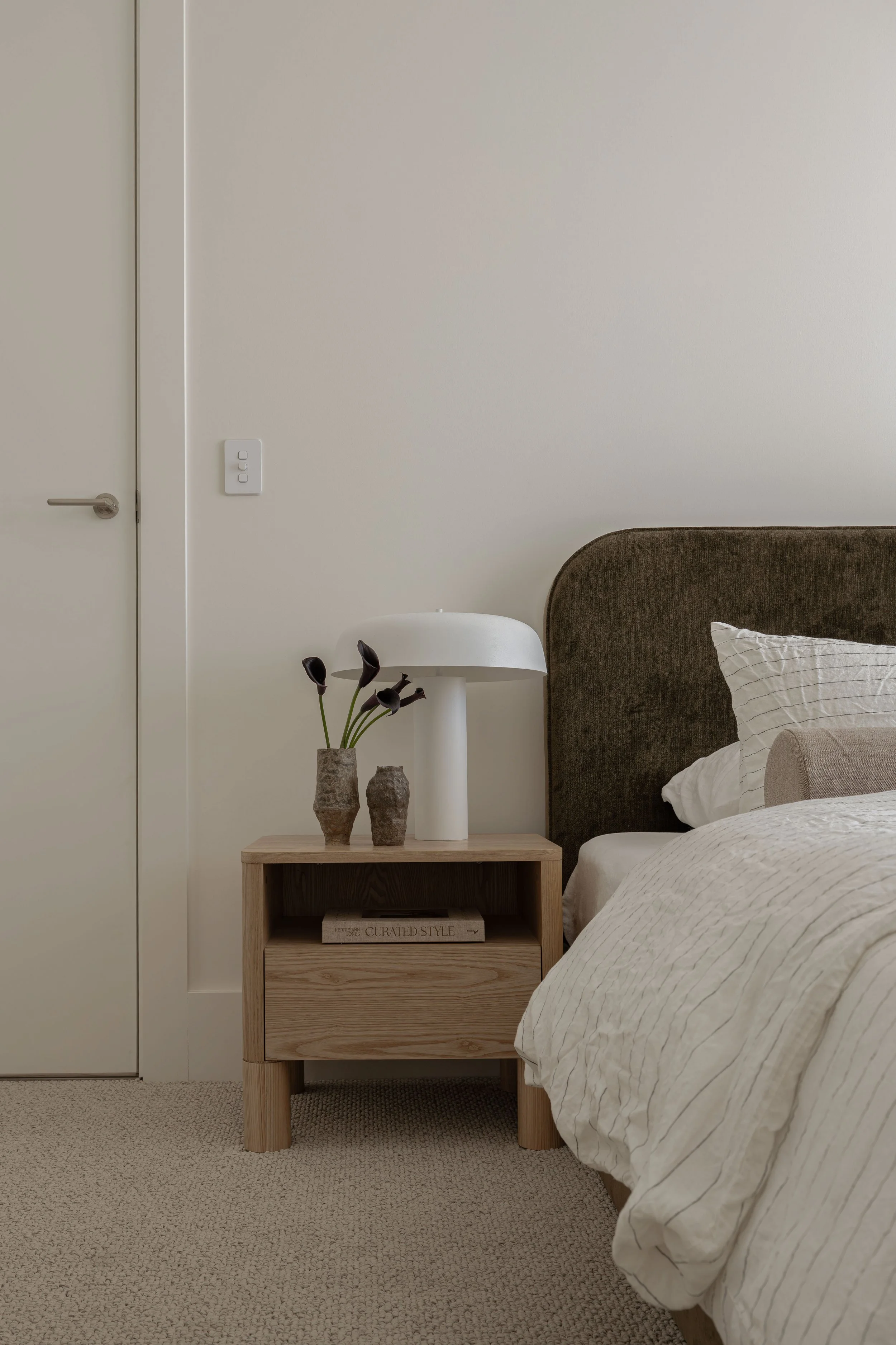 A minimalist bedroom with a beige carpet, a wooden nightstand with a drawer and shelf, a white modern table lamp, decorative vases with dark flowers, a bed with white striped bedding, a dark brown upholstered headboard, a white pillow with a black grid pattern, a white wall, a light switch, and a closed door.