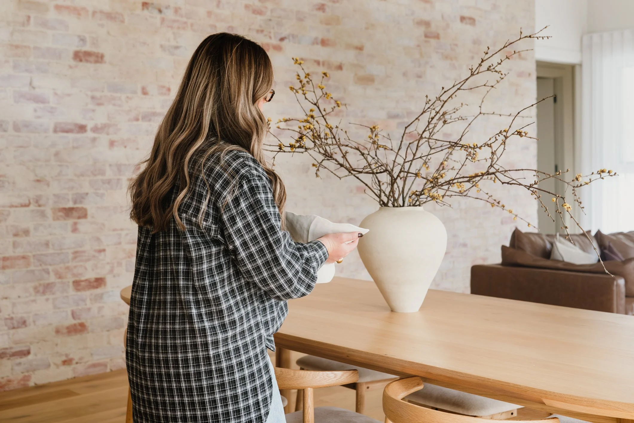 A woman with long wavy hair and glasses, wearing a checkered blazer, is arranging a ceramic vase with branches and yellow flowers on a wooden table in a bright room with a brick wall and a window.