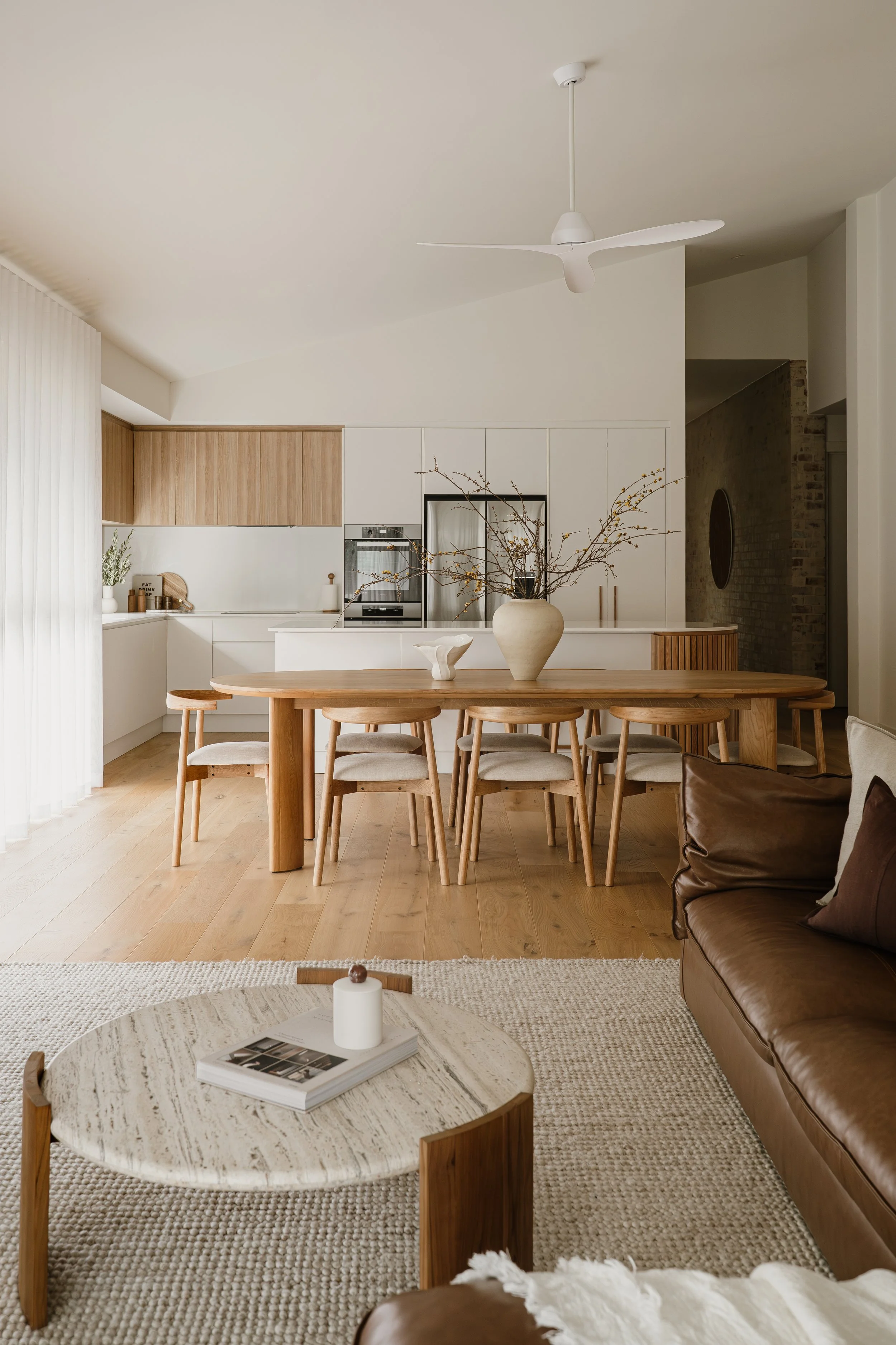 A modern, minimalist kitchen and dining area with a light wood dining table, matching chairs, a large vase with branches on the table, and a brown leather sofa with a pillow in the foreground. The background features a white and wood kitchen with a built-in oven, a ceiling fan, and a brick wall partially visible to the right.