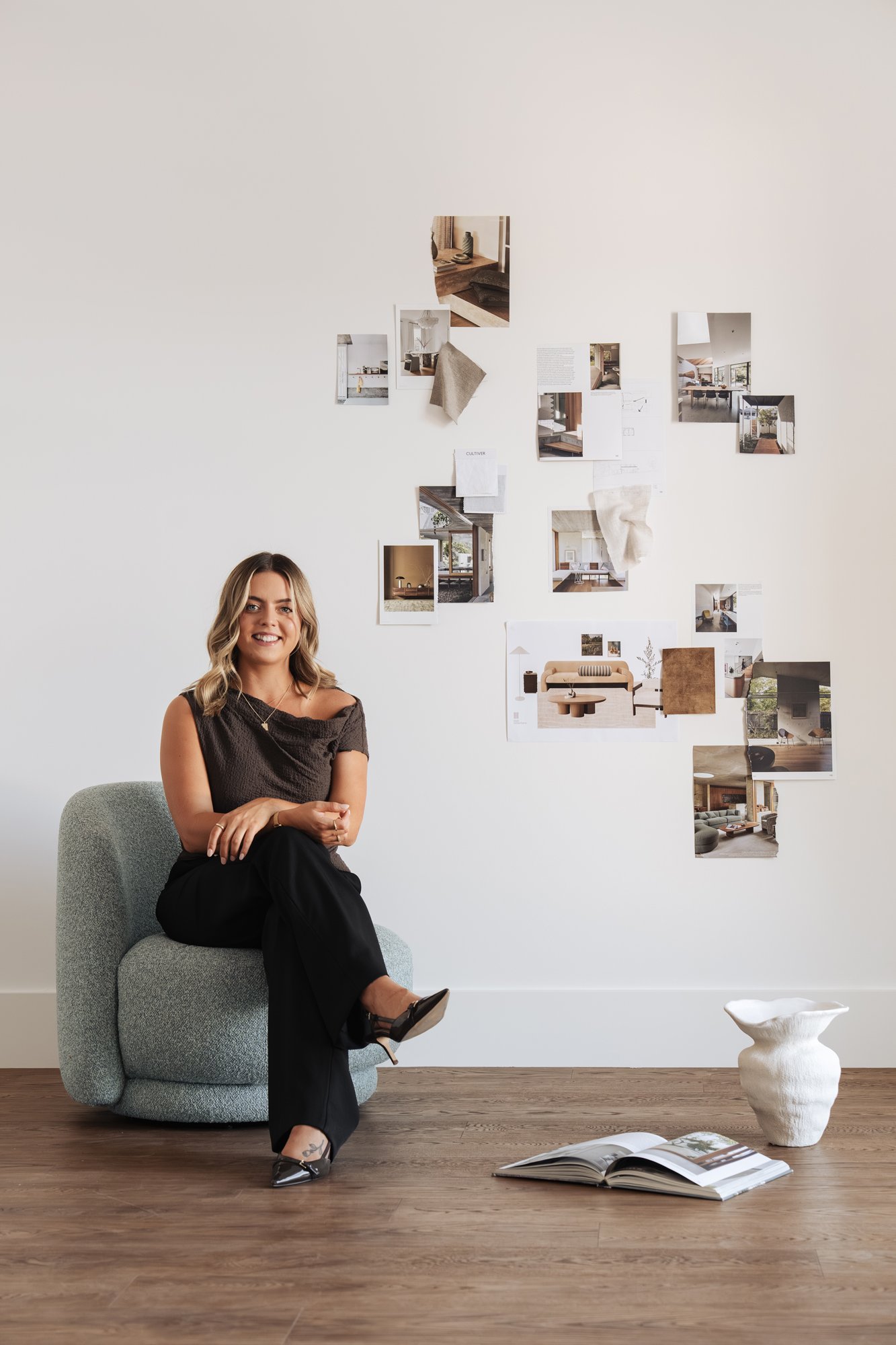 A woman sitting on a light gray, textured modern armchair in a minimalist room with a wooden floor, white wall with photos, a coffee table, and an open magazine.
