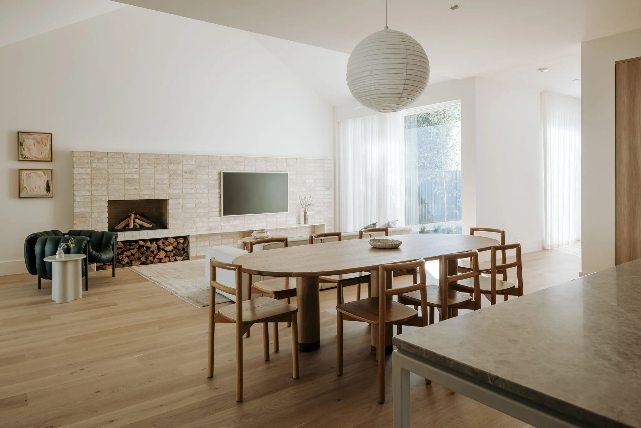 A bright, modern living and dining area with a large wooden dining table, surrounding chairs, a white fireplace with stacked logs, a black armchair, a side table, and a wall-mounted television. Floor-to-ceiling windows with white curtains allow natural light to fill the space.