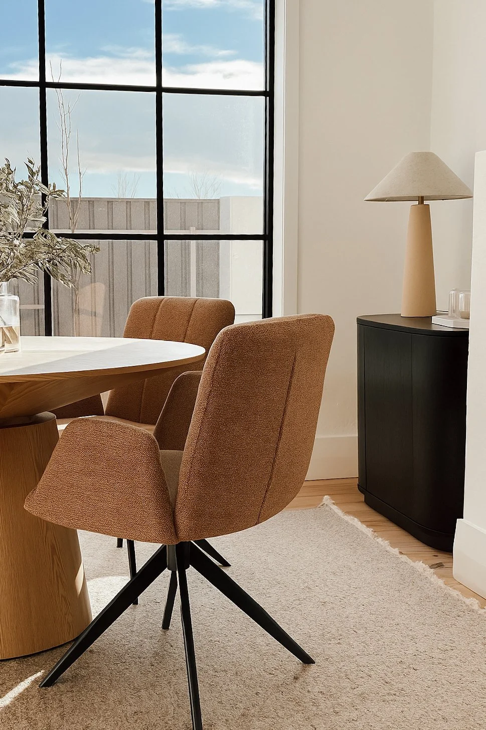 A cozy corner of a dining area with a round wooden table, two upholstered chairs in a warm terracotta color, a black cabinet with a beige table lamp on top, a beige rug on light wood flooring, and a large window with black frame showing trees and a partly cloudy sky outside.