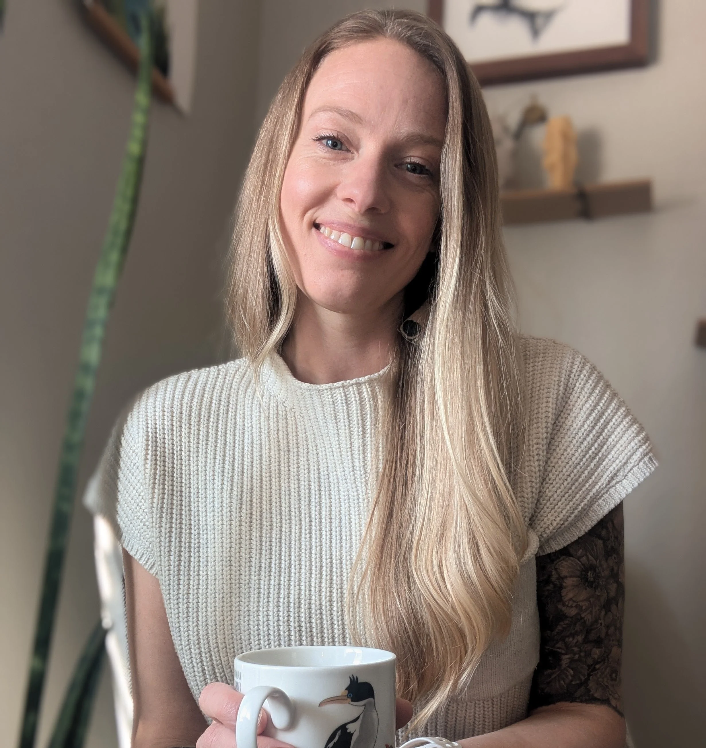 A woman with long blonde hair smiling, holding a white coffee mug with a black penguin illustration, wearing a beige sweater, sitting indoors near a light-colored wall with framed artwork.