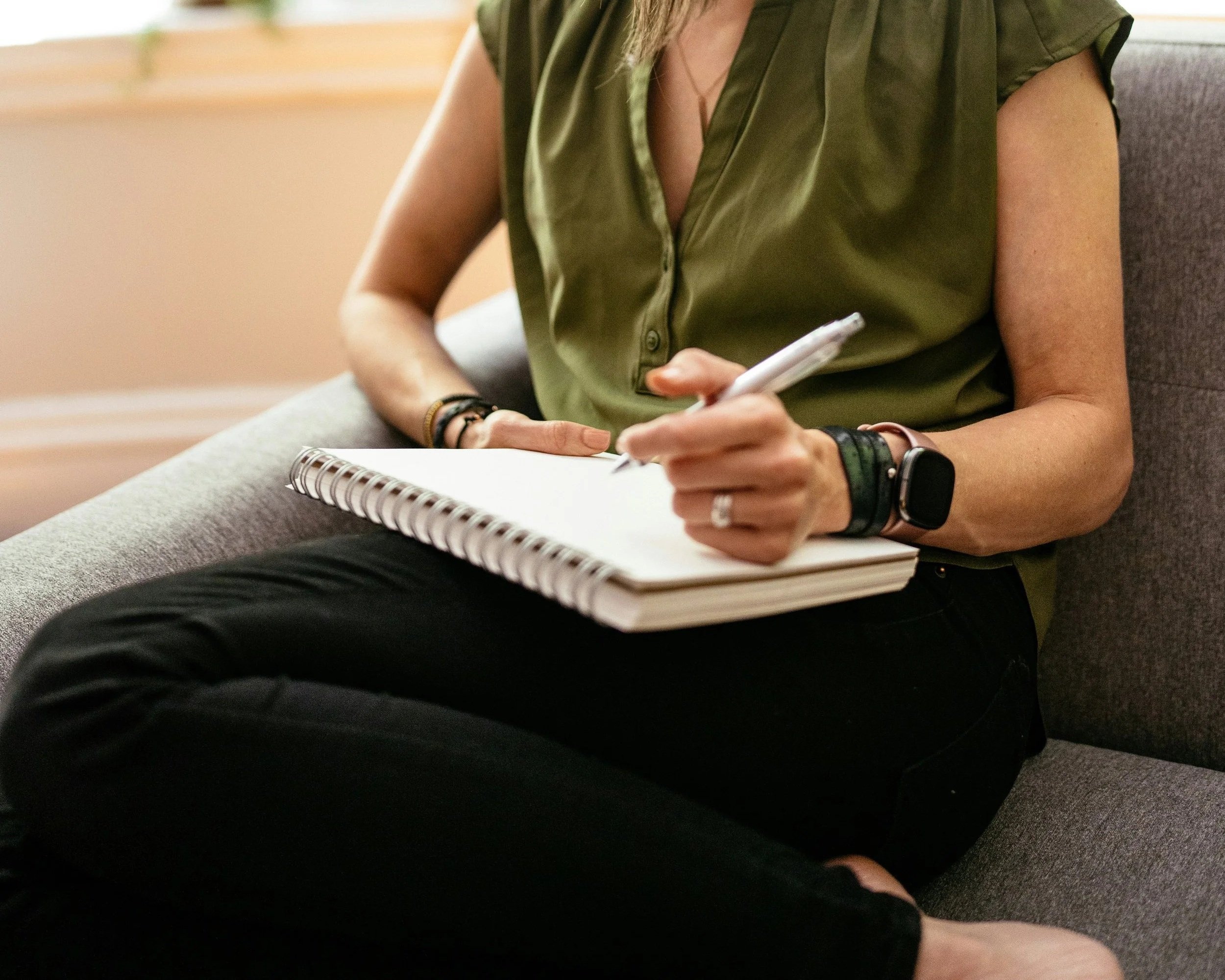 A woman wearing a gold ring, bracelets, and a smartwatch, sitting on a gray couch. She is holding a pen in her right hand and a spiral notebook on her lap. She is wearing a green, sleeveless, button-up shirt with her left hand resting on her leg. The background is blurred.