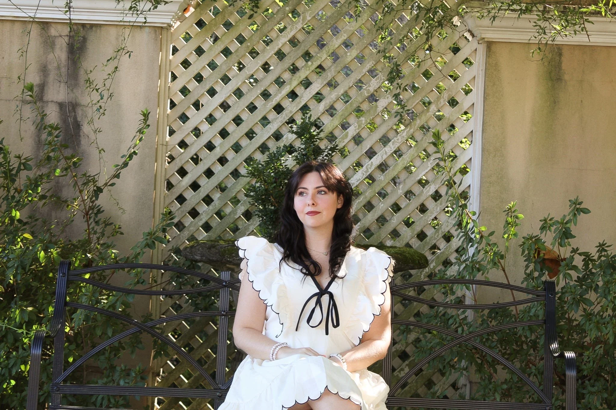 A woman with black hair and light skin sitting on a black metal bench in a garden with green plants and a lattice fence, wearing a white dress with black trim, looking to her right.