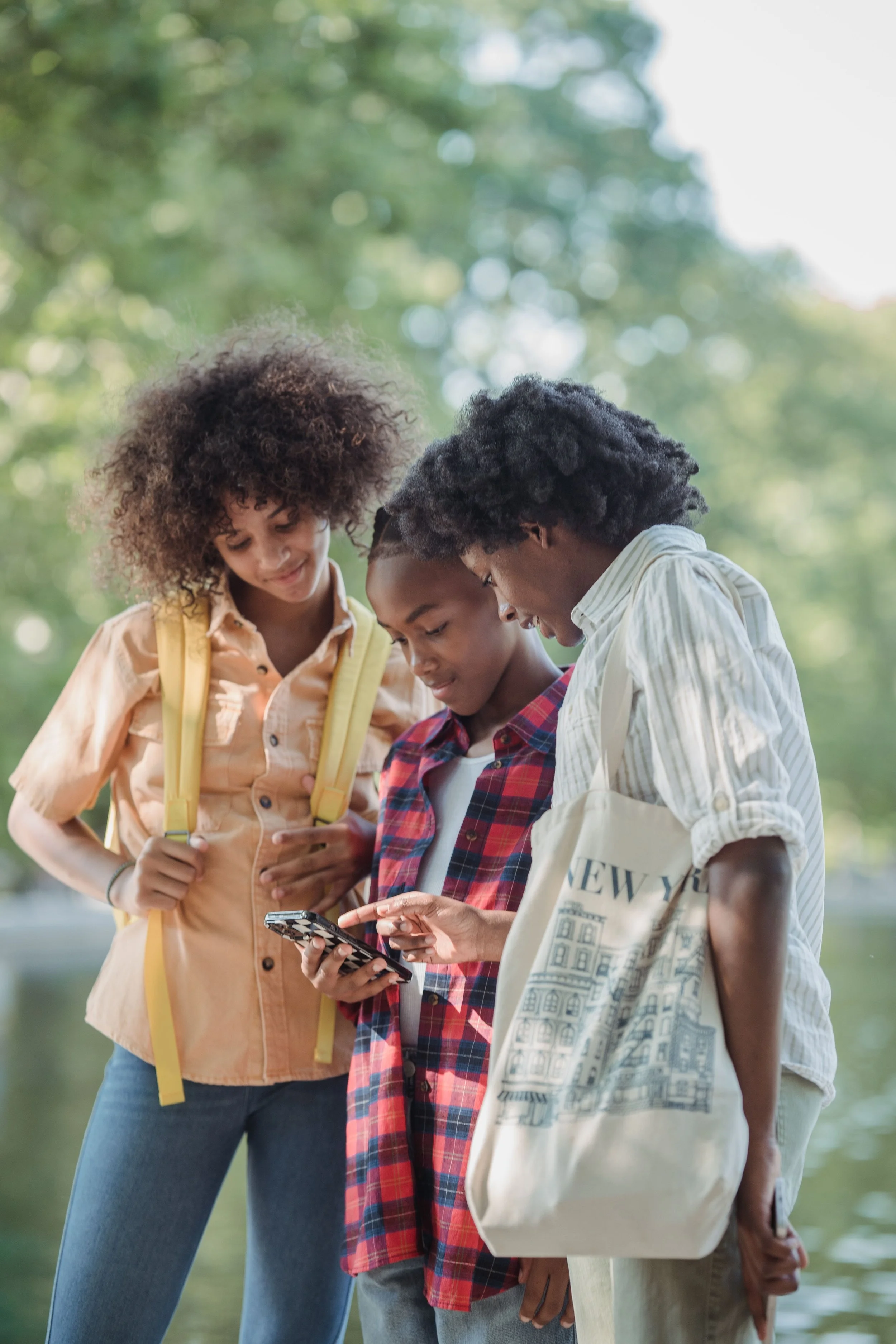 Three teenagers, two girls and one boy, look at a smartphone outdoors near a body of water, with green trees in the background.