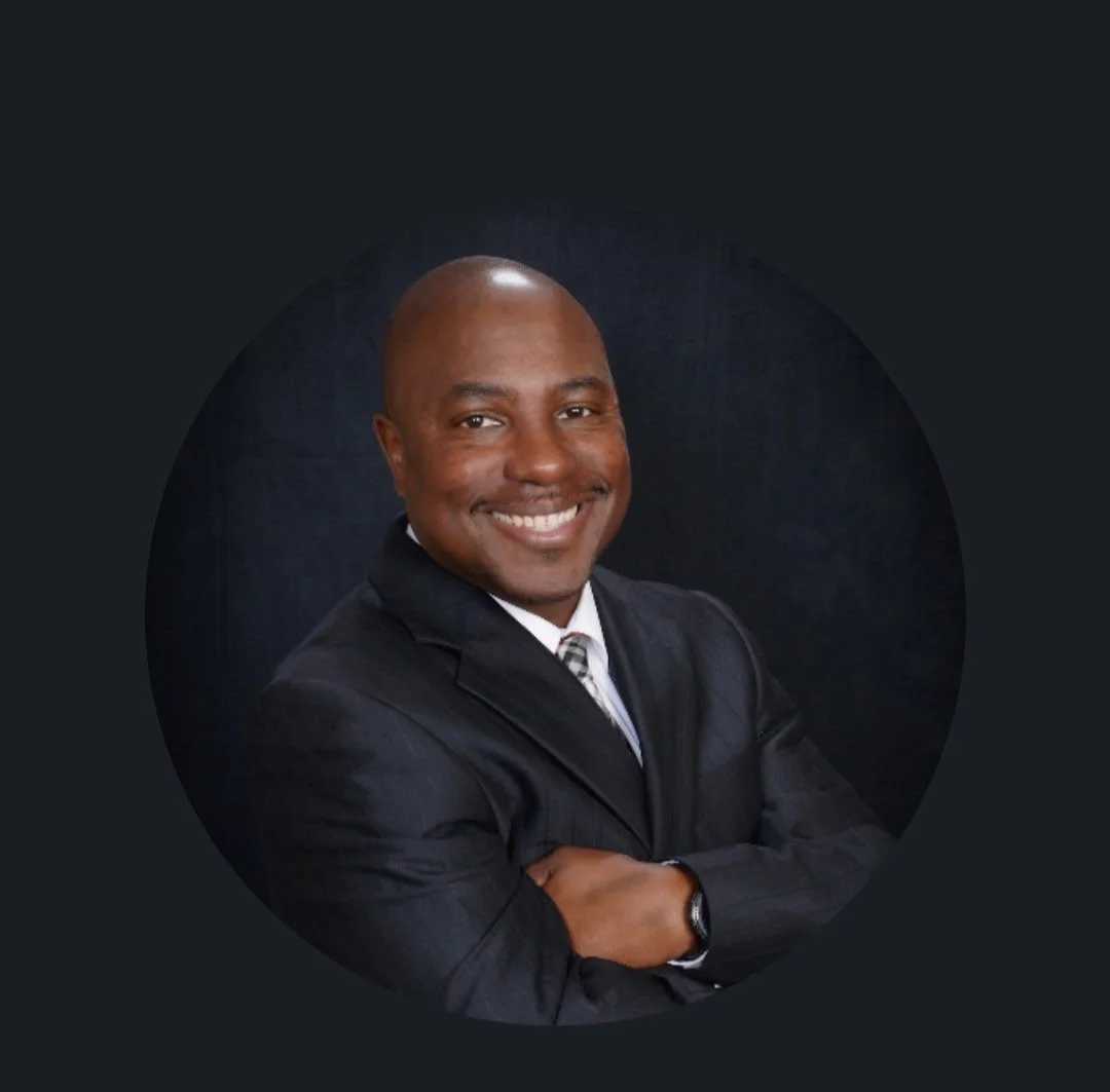 Professional headshot of a smiling man in a black suit, crossed arms, dark background.