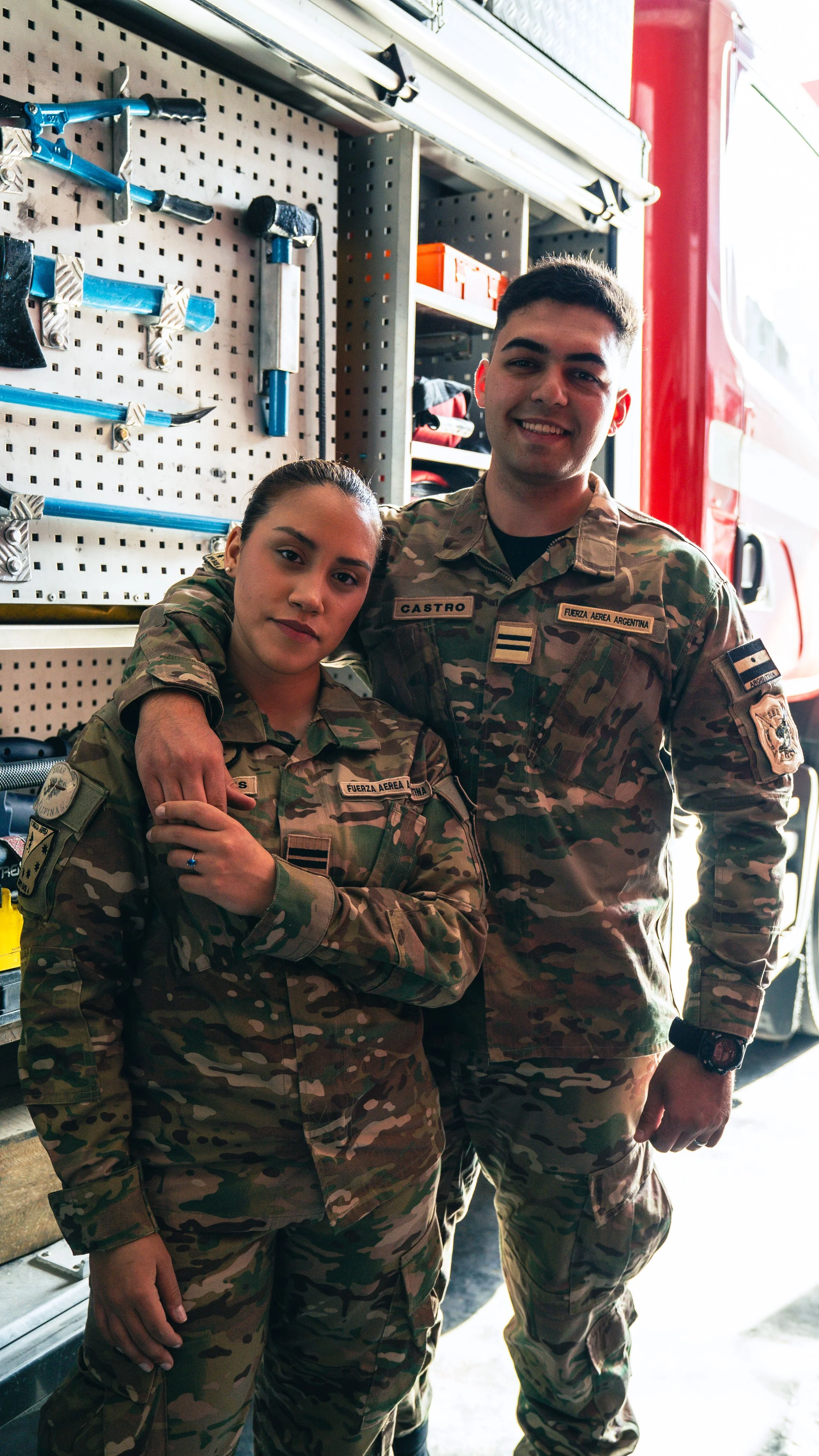 Two soldiers in camouflage uniforms standing inside a fire station, smiling at the camera. One has her arm around the other's shoulder. Both have patches and name tags on their uniforms.