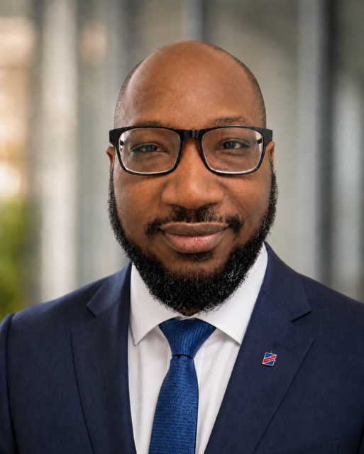 Professional man with glasses, beard, wearing a navy suit, white shirt, and blue tie, smiling outdoors.