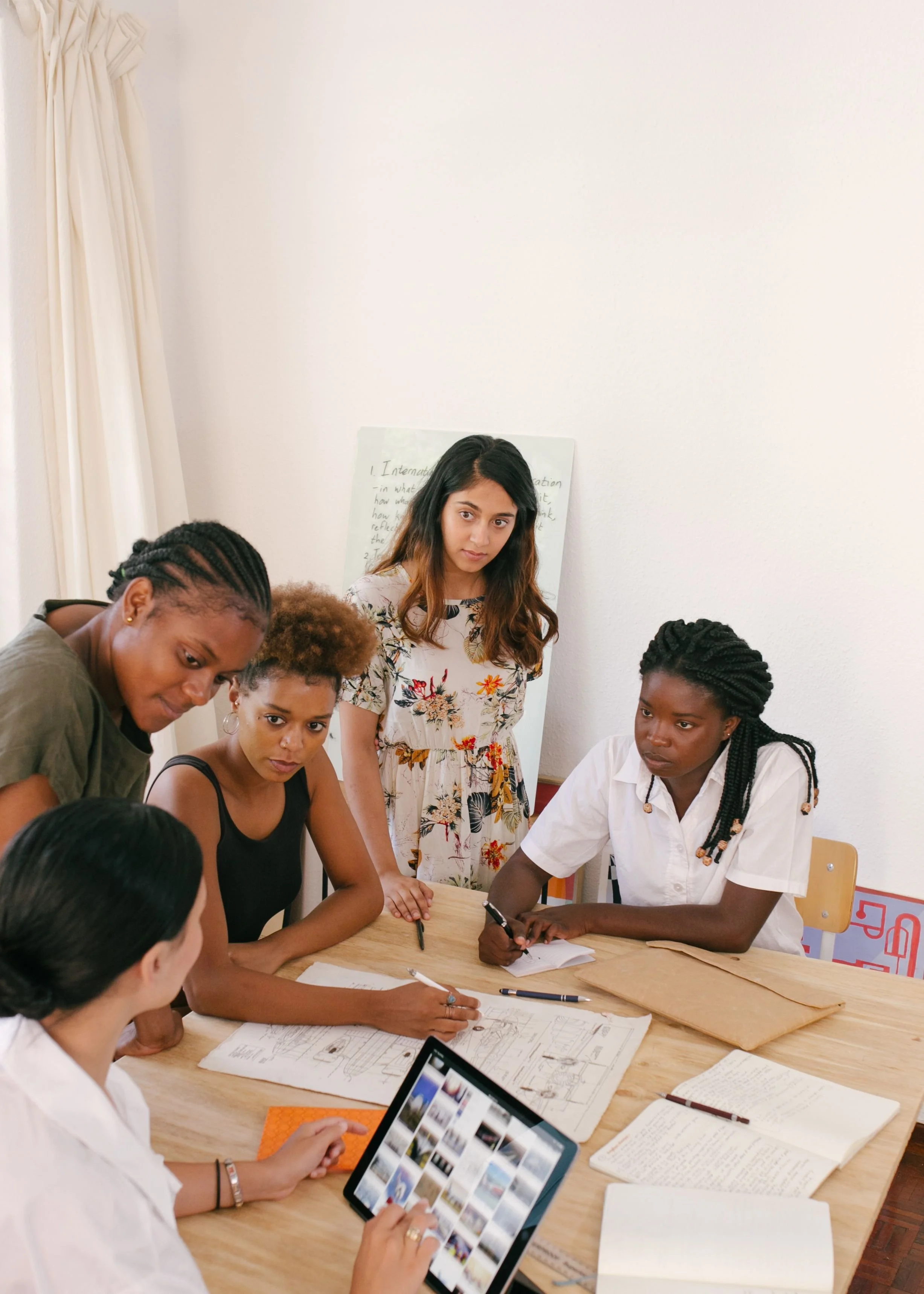 A diverse group of women gathered around a table, engaged in a discussion or collaborative work in a bright room with white walls, some taking notes, a woman in a white floral dress standing, and a woman holding a tablet displaying multiple images.