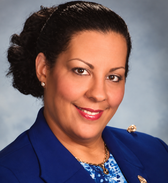 A woman with dark hair styled in an updo, wearing a blue blazer and a patterned blue blouse, smiling with red lipstick, earrings, and a necklace, against a gray background.